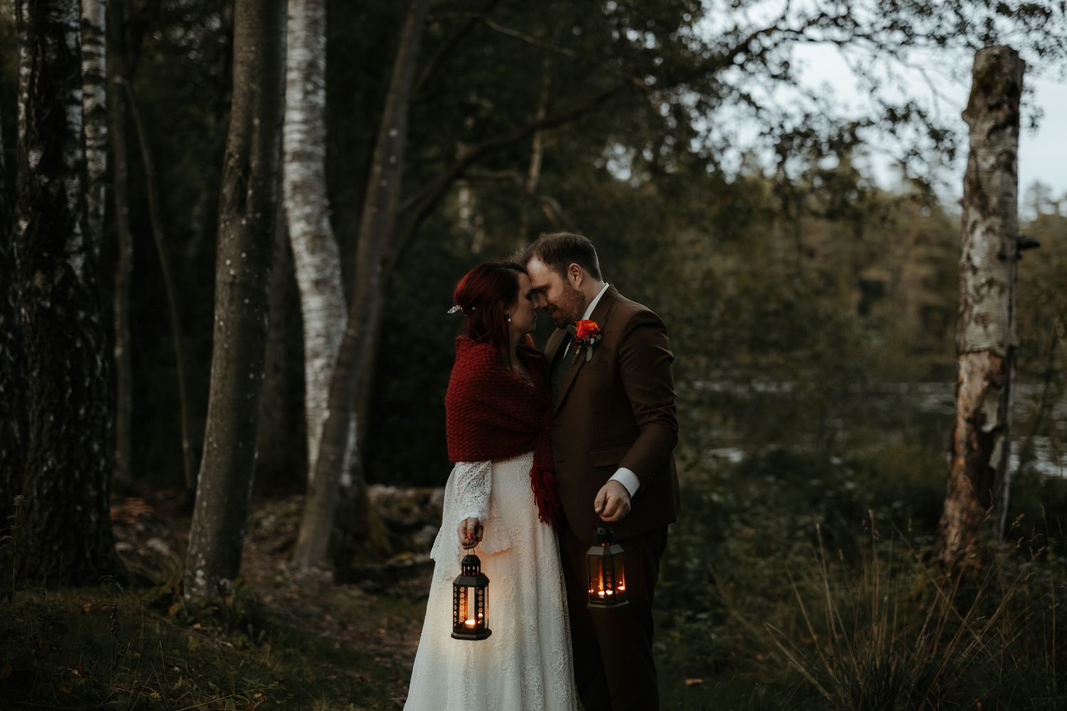 Brudpar står panna mot panna framför en sjö i skymningsljus med en varsin lykta i handen. Foto av Grevholm Photography.