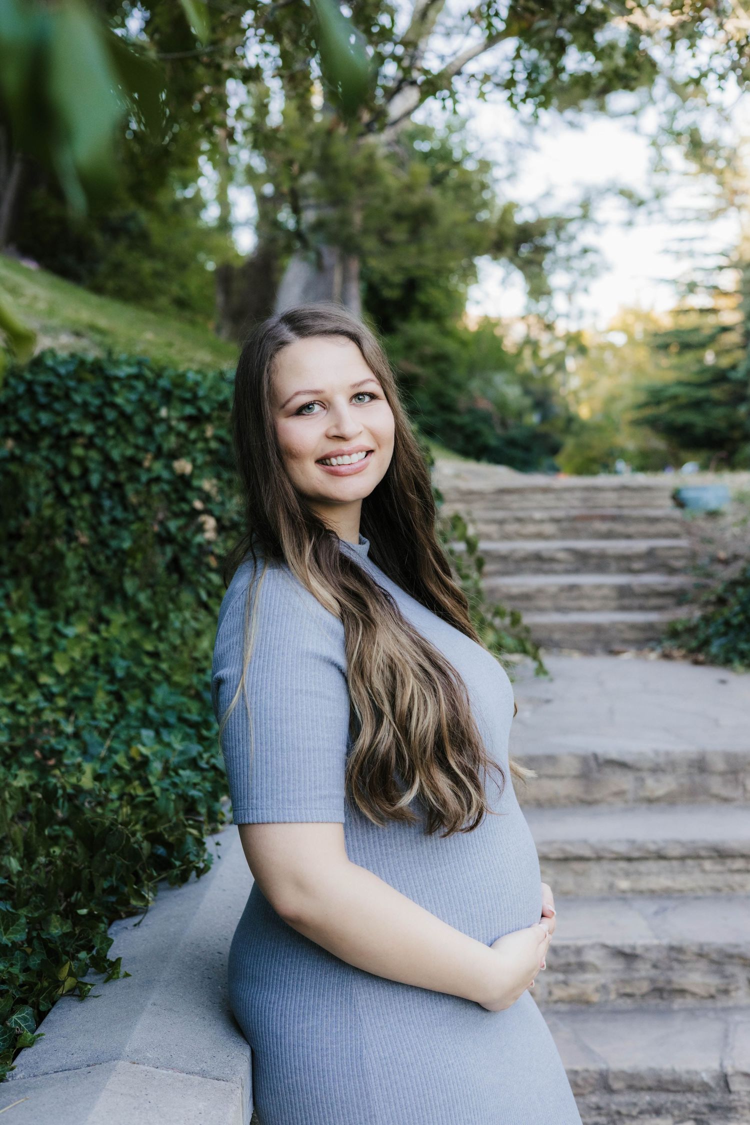 Expectant mother in gray dress poses on stone steps surrounded by greenery.