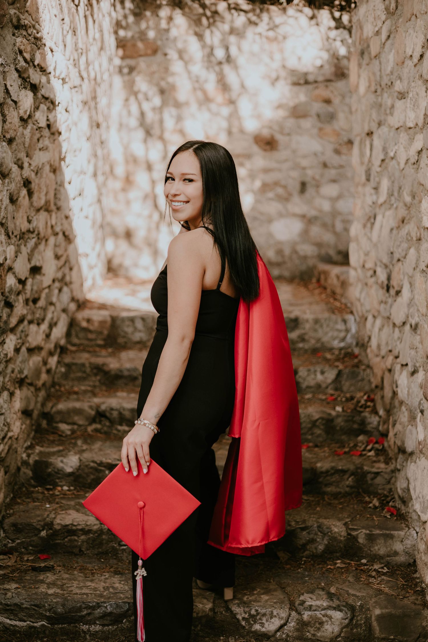 A graduate in black dress and red graduation gown poses against stone walls.