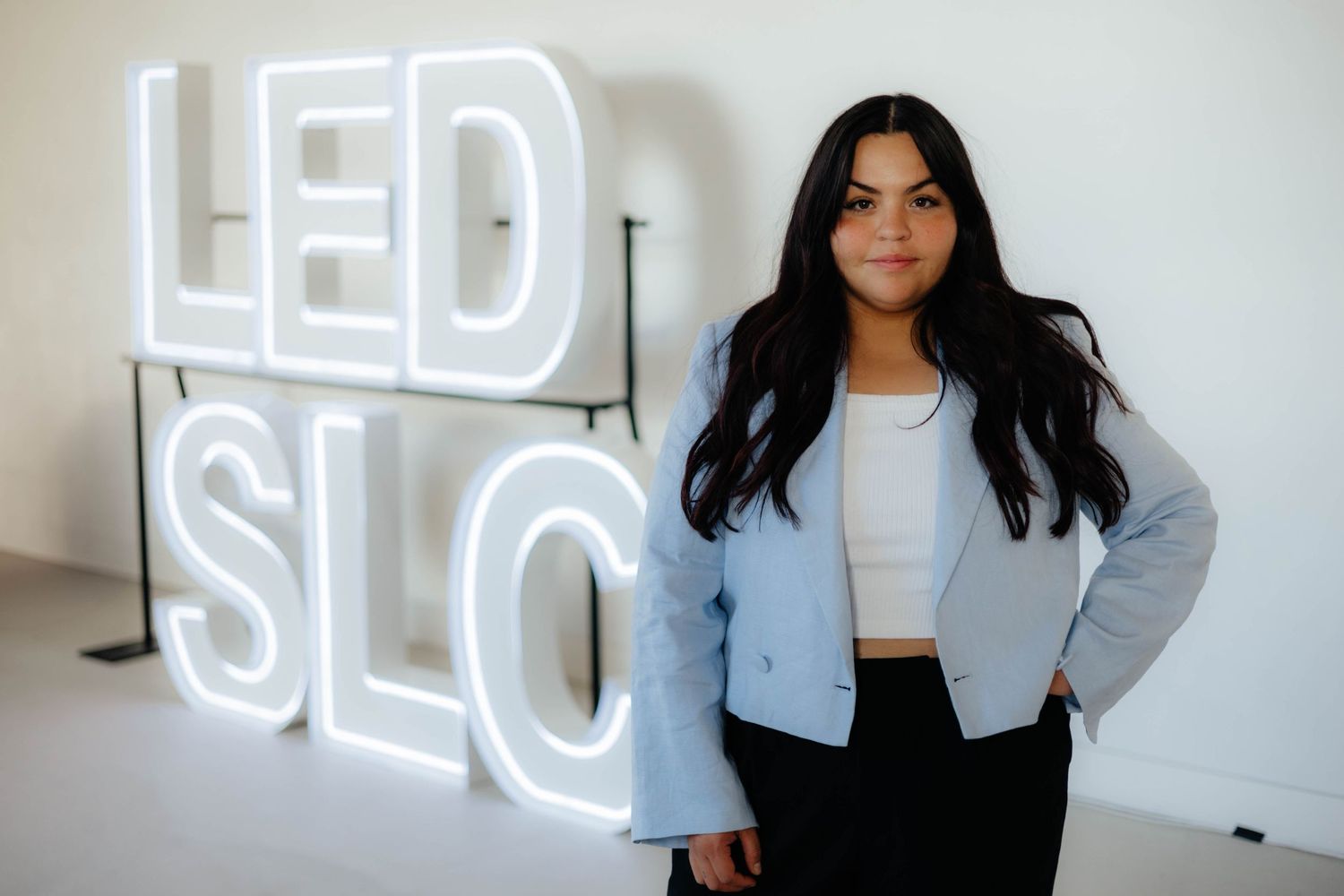 Professional stands in front of illuminated LED SLO sign wearing light blue blazer and white crop top.