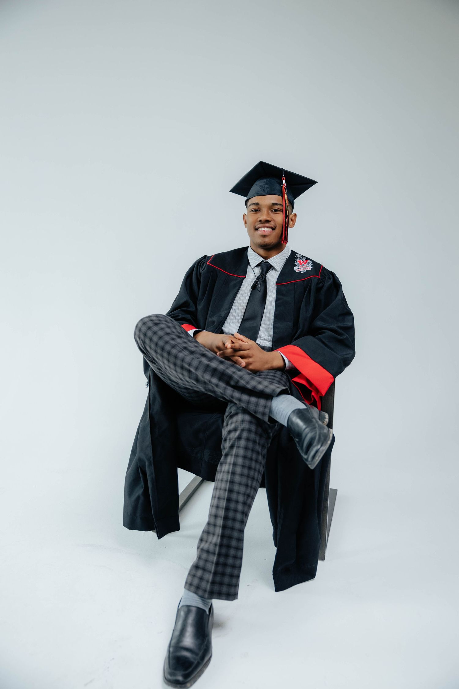 Graduate in black cap and gown sits confidently wearing checkered pants and dress shoes.