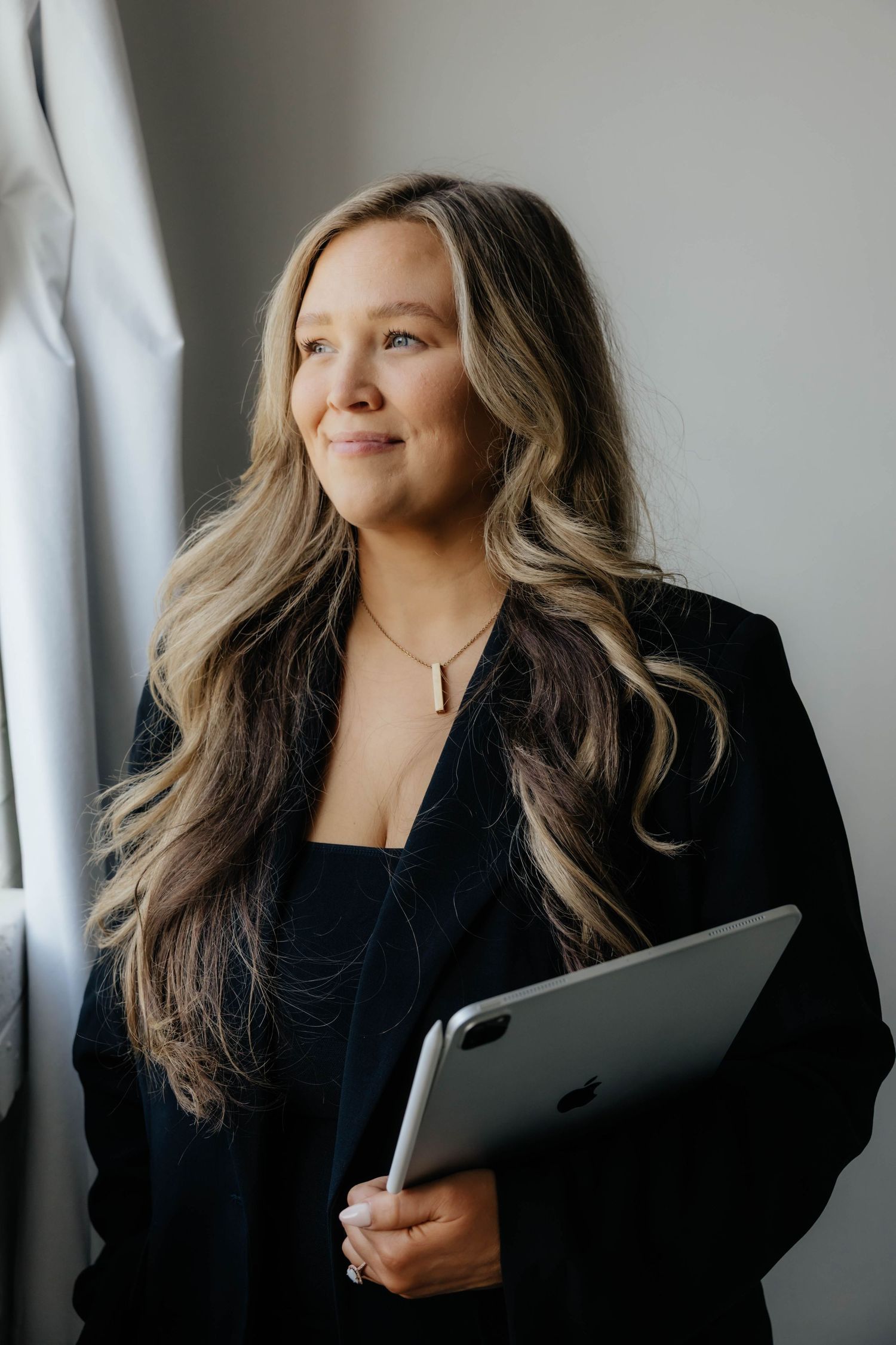 Professional woman in black blazer poses confidently with laptop in modern setting.