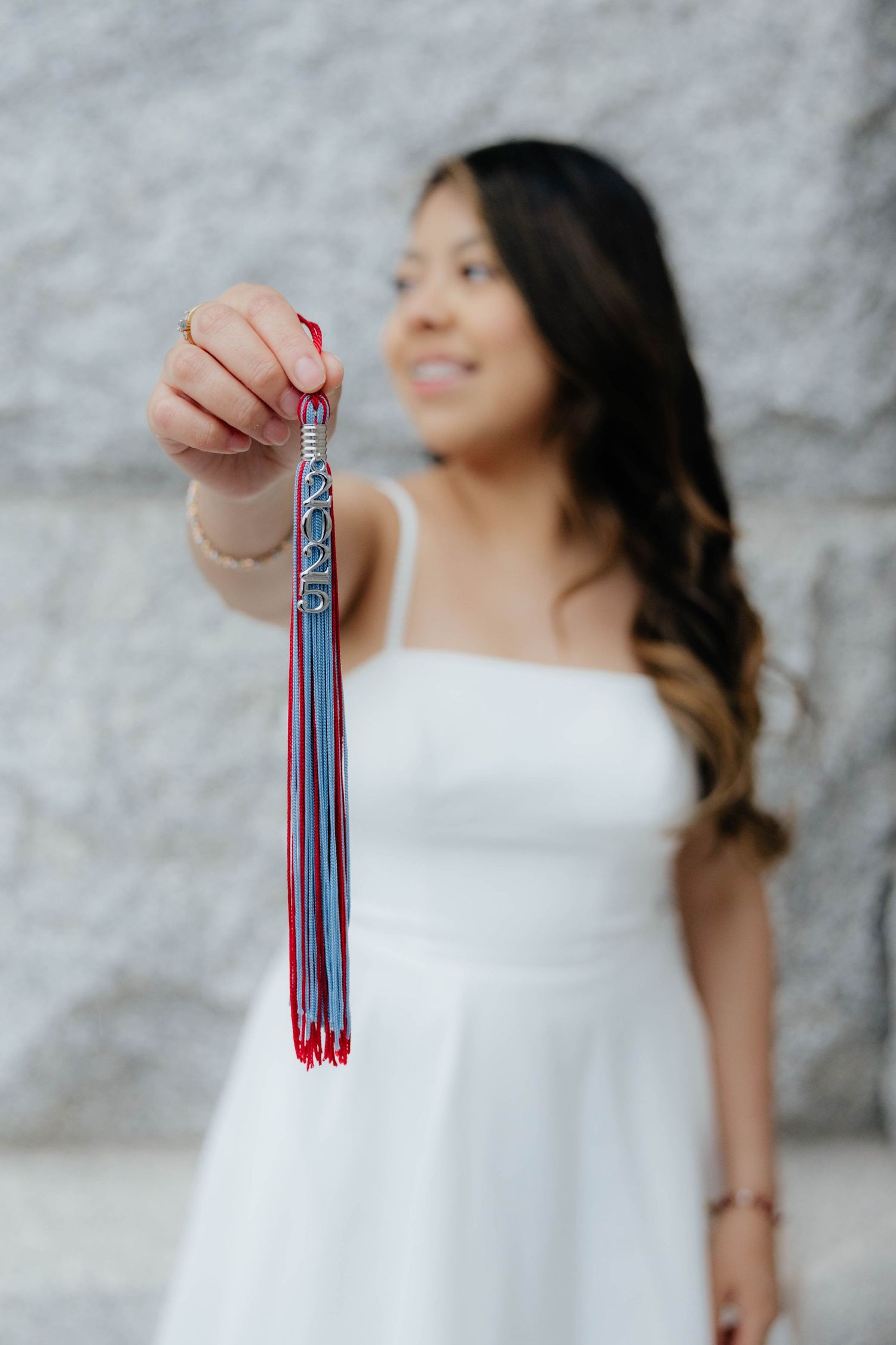 Graduate holds red and blue tassel while wearing white dress against stone wall.