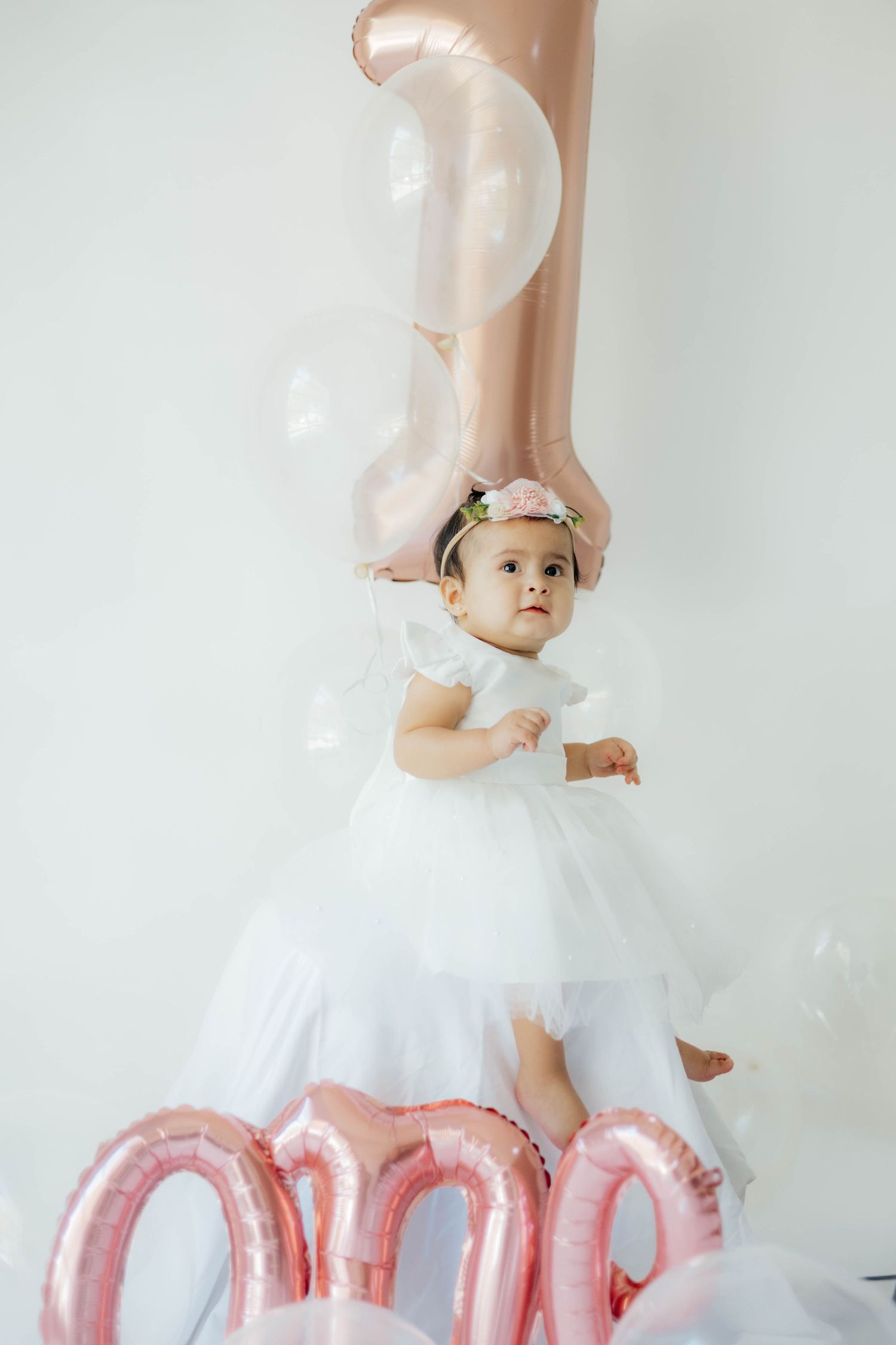 Baby photography with rose gold balloons and white tulle dress against light background.