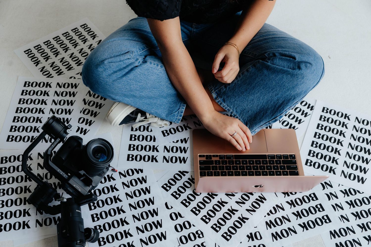 Overhead view of someone working with camera and laptop on patterned 'Book Now' paper.