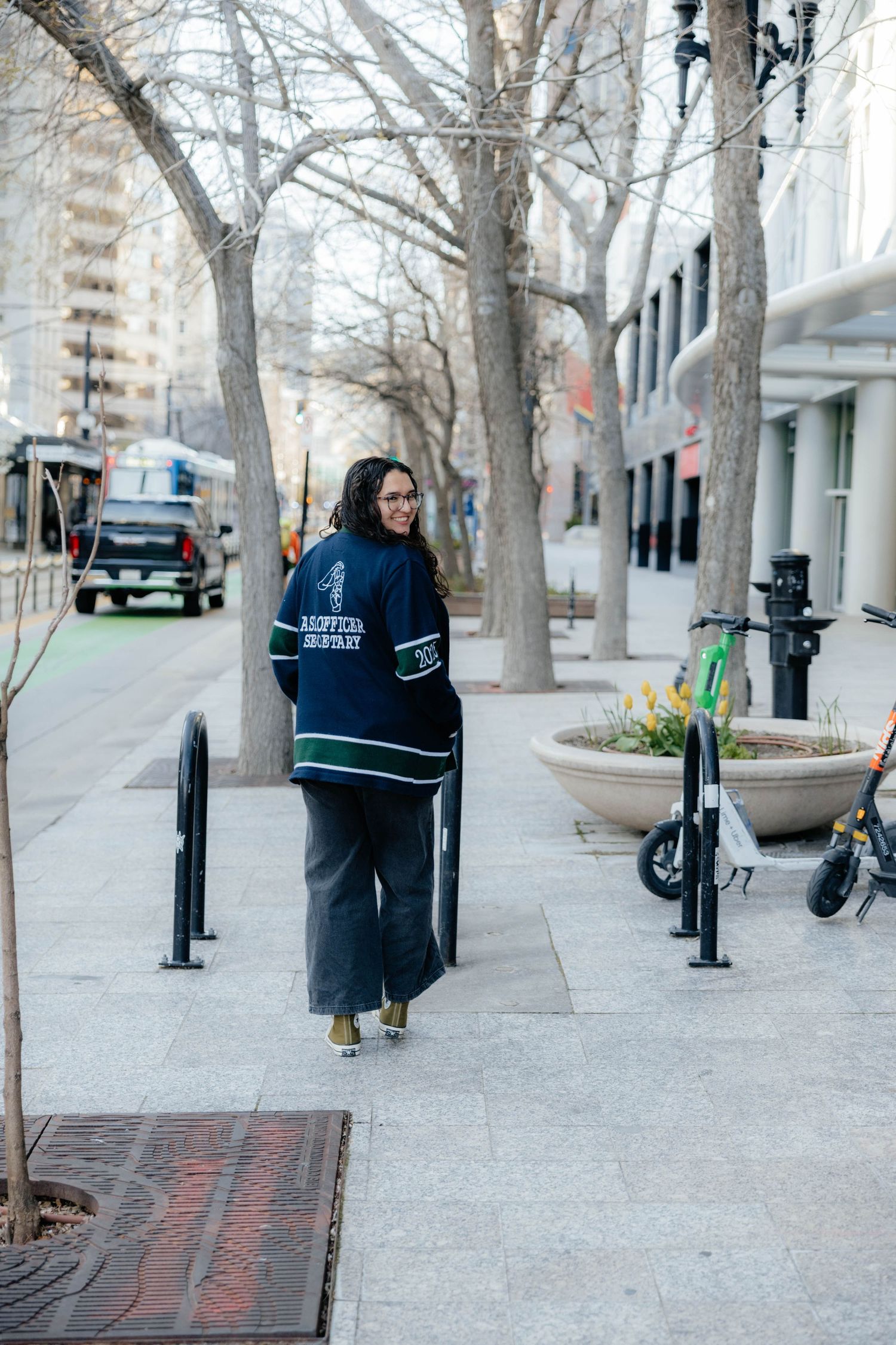 Student walks down city sidewalk wearing dark sweater on winter day with bare trees.