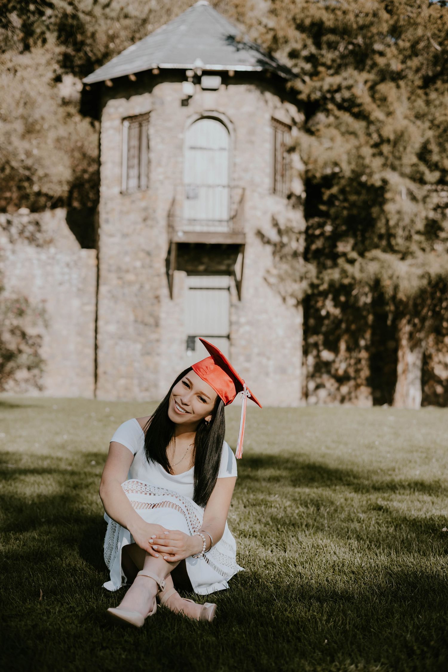 Graduate in red cap poses on grass with historic stone building in background.