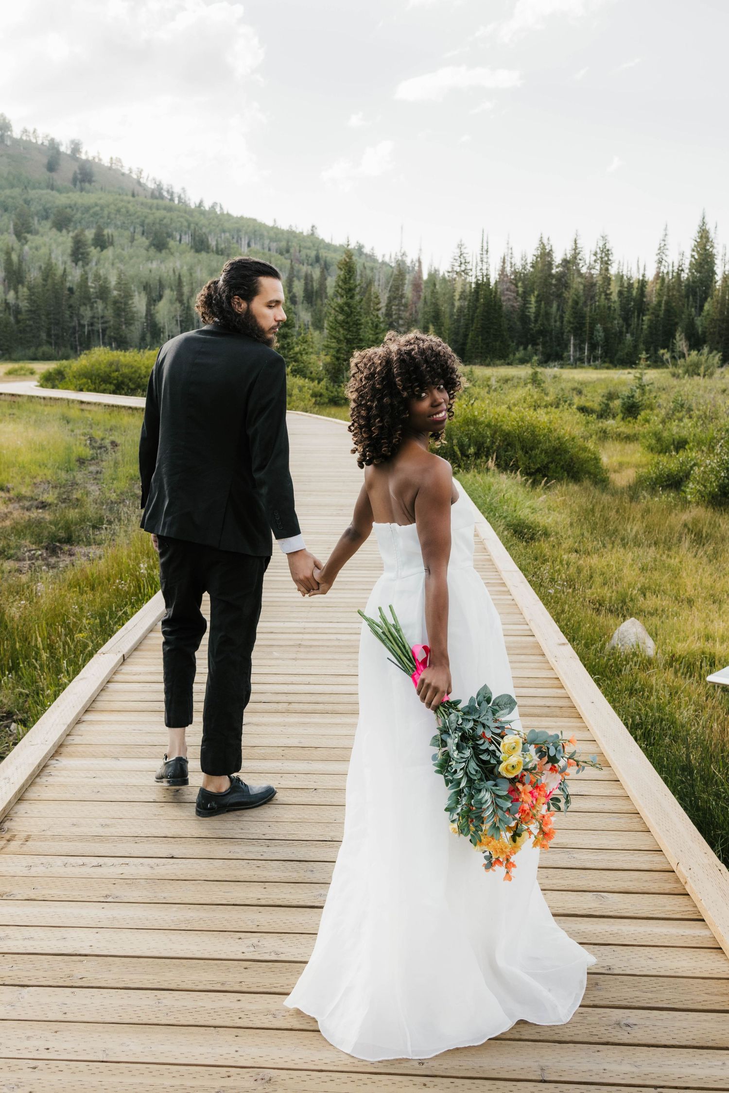 Couple walks hand in hand on a wooden boardwalk through mountain scenery.