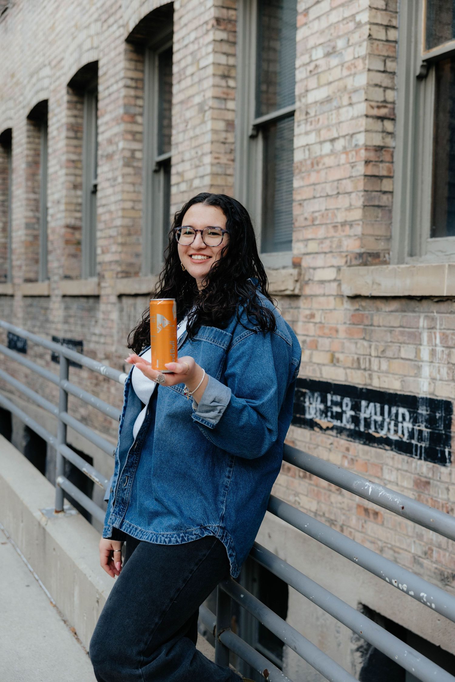 Student in denim jacket holds orange drink while standing against brick building.