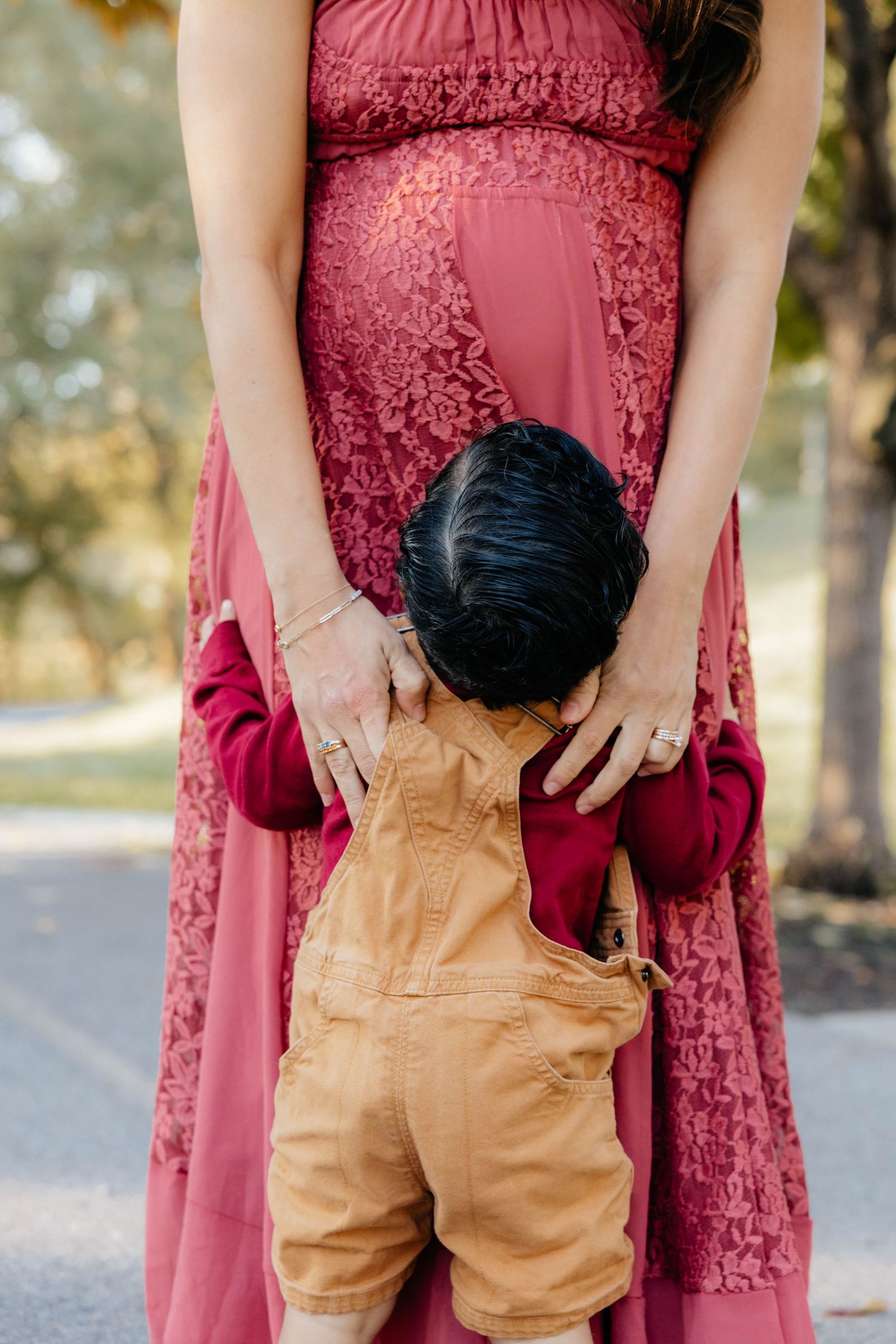 Small brown overalls held up against a burgundy maternity dress in an outdoor setting.