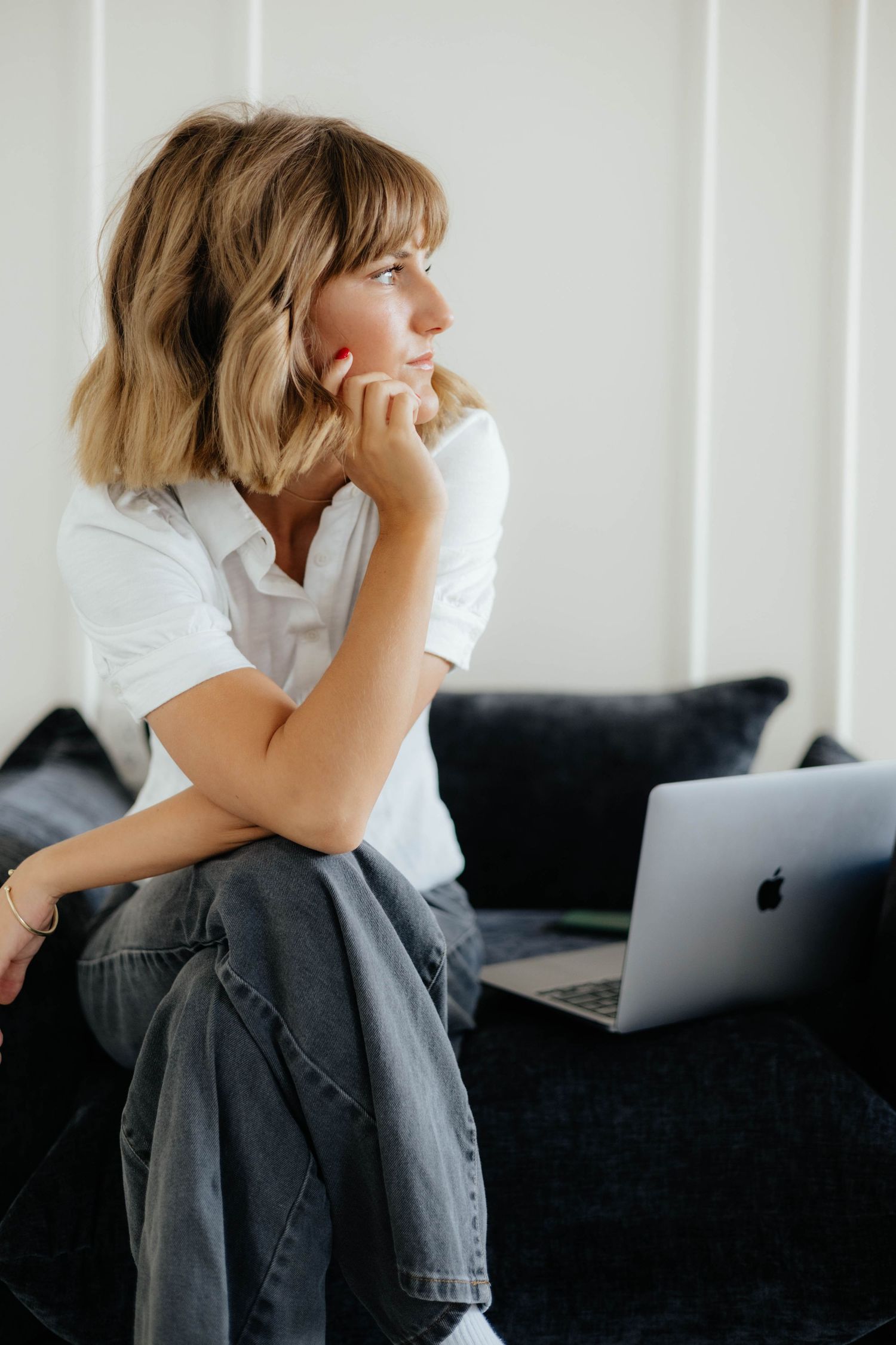 Woman in white shirt working on MacBook while sitting thoughtfully on dark couch.