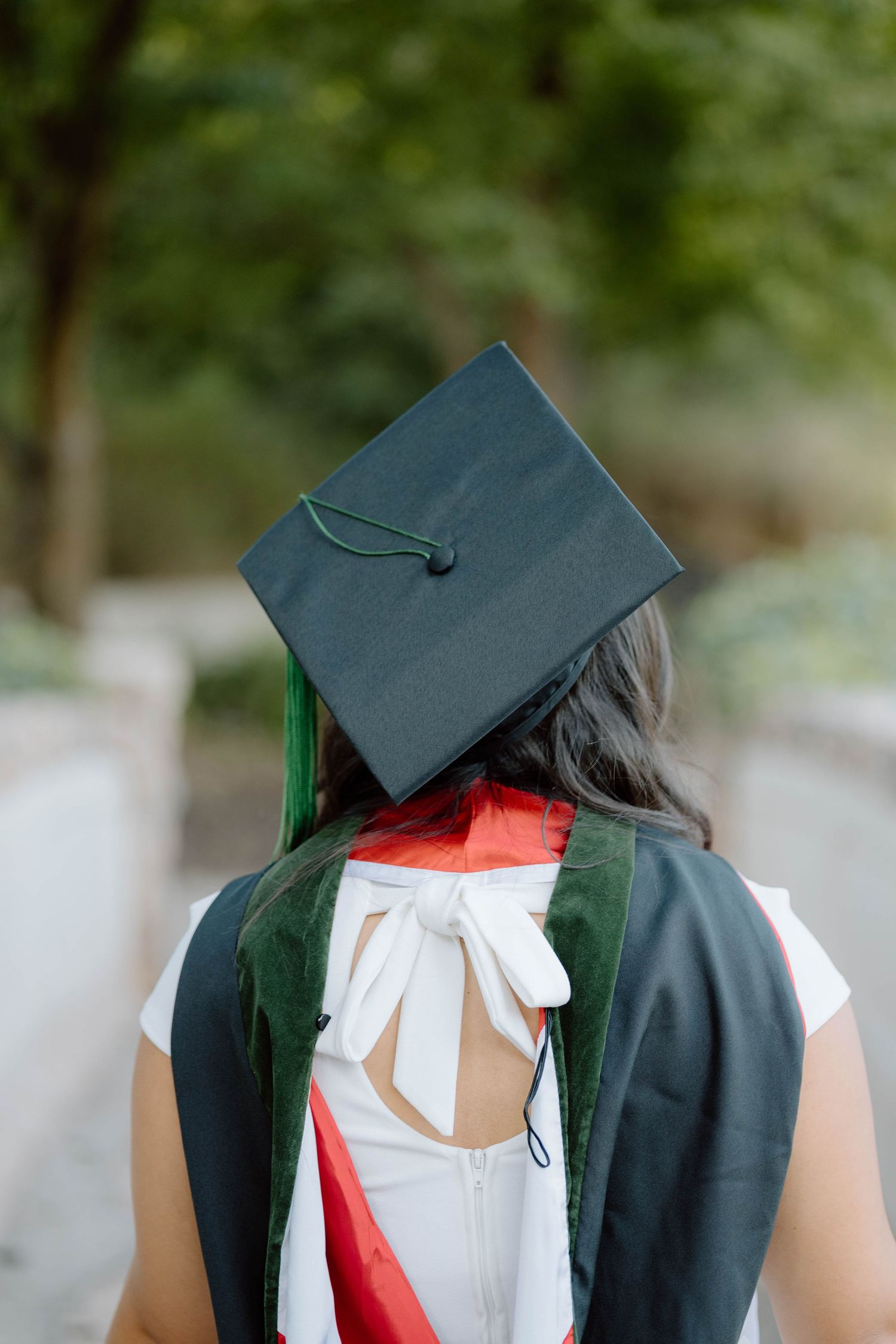 Graduate wearing dark cap and gown with white dress and red sash seen from behind.