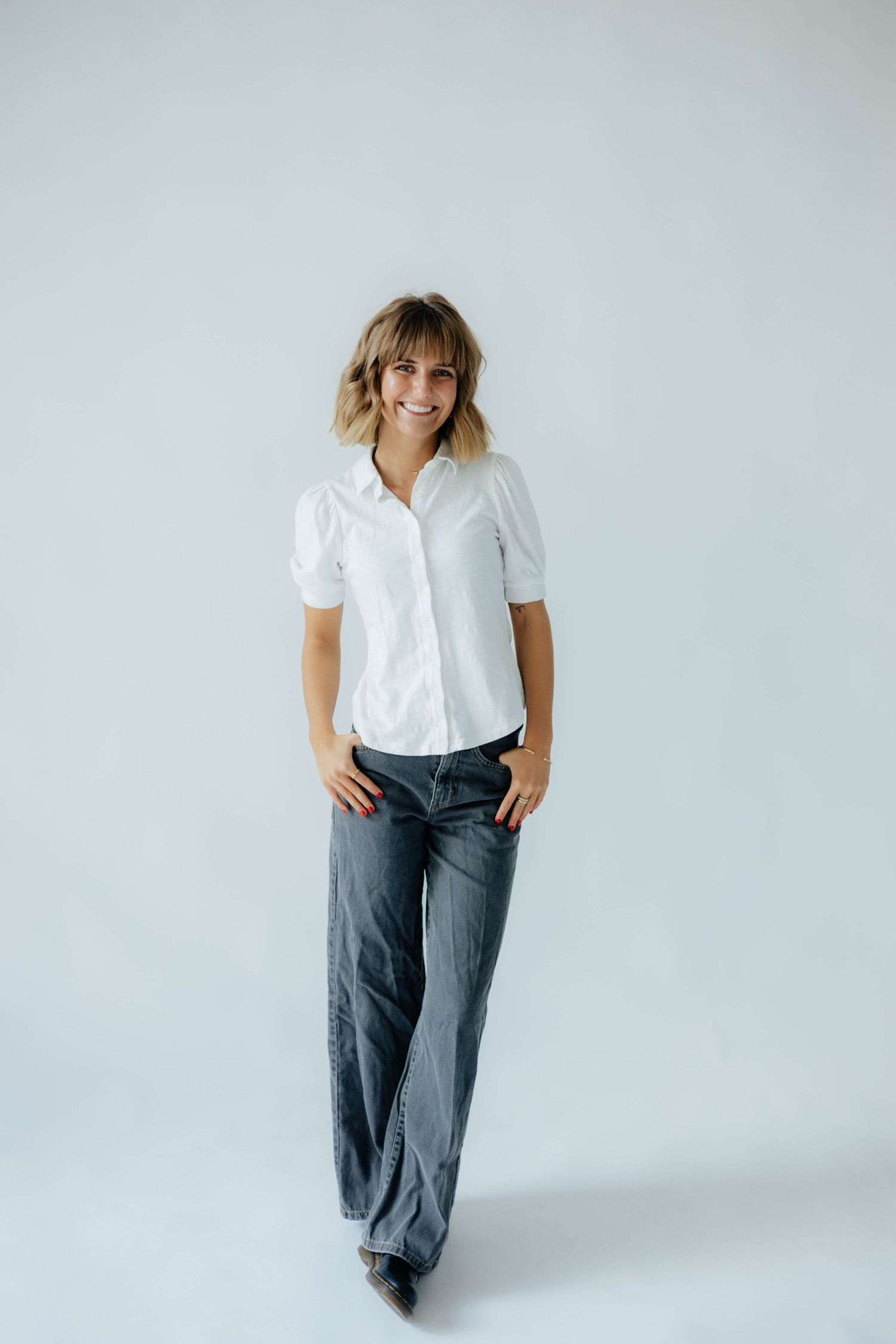 Woman in white button-up shirt and gray pants poses against white backdrop in studio setting.