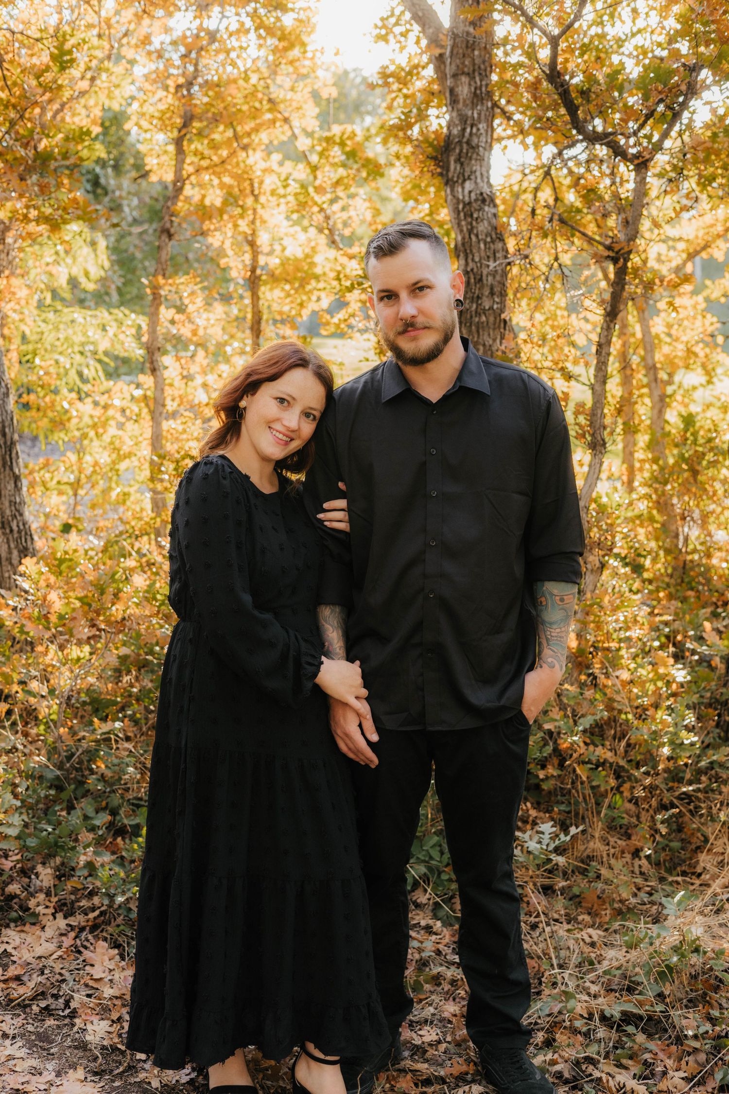 Couple in matching black outfits pose together in golden autumn forest.