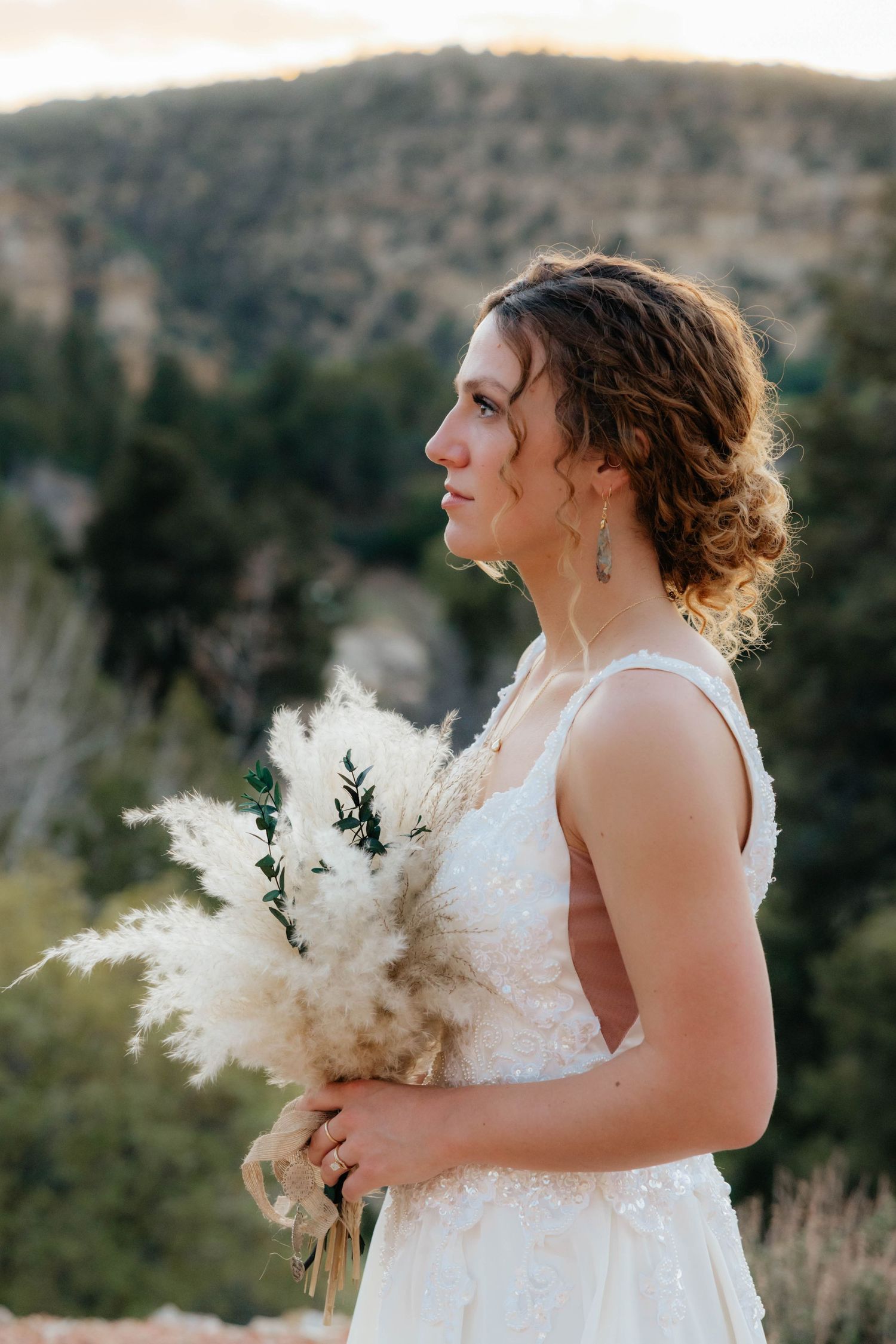 A bride in a white lace dress holds a dried pampas grass bouquet while standing against a mountainous backdrop at sunset.