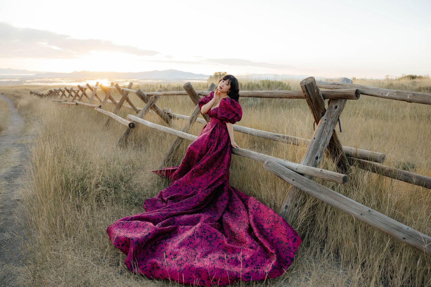 Dramatic pose in burgundy gown against rustic wooden fence at sunset.