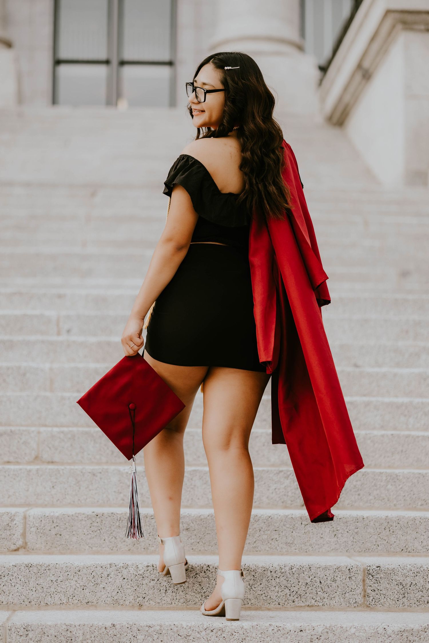 A graduate in a black dress and red graduation gown poses on stone steps.