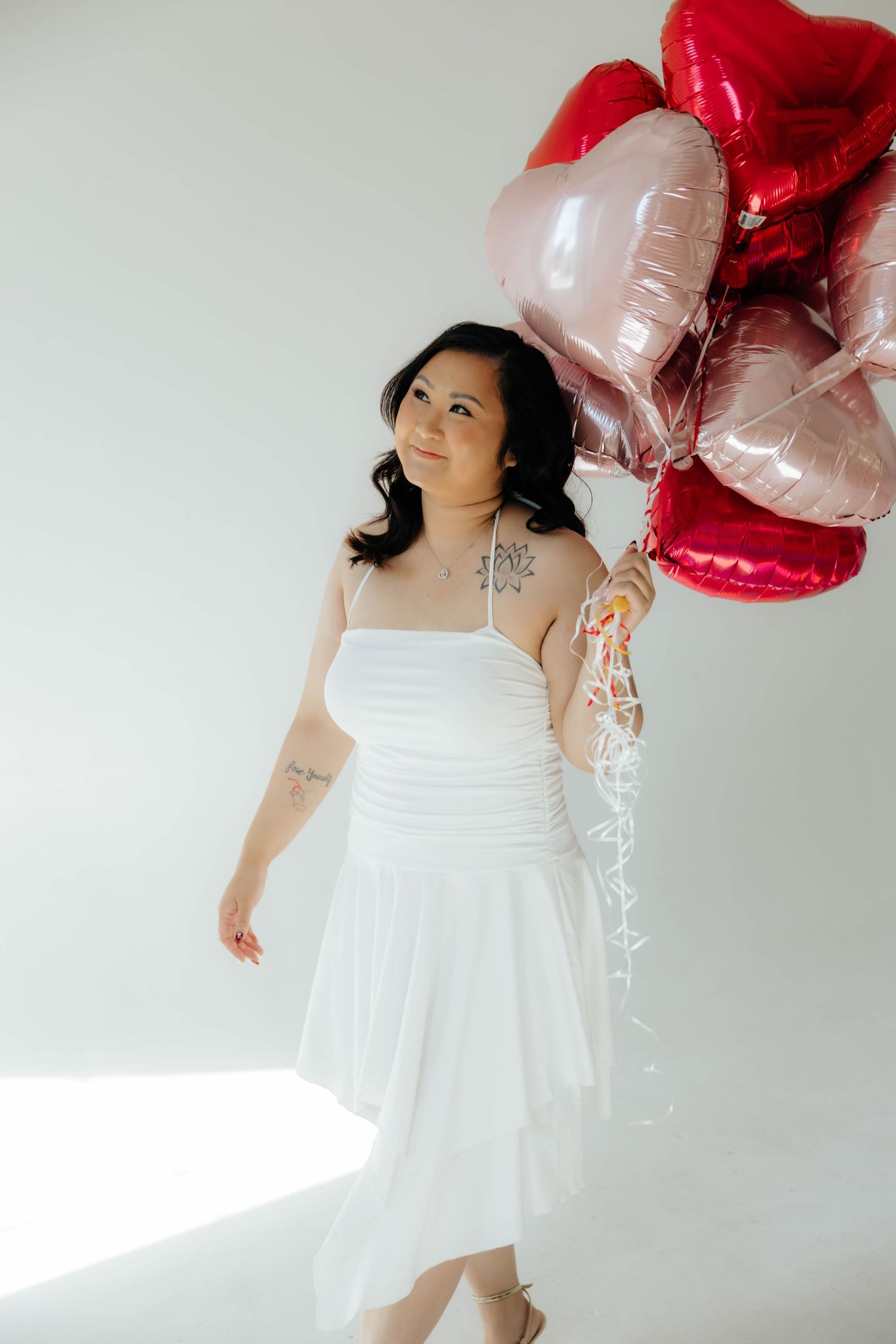 Woman in white dress poses elegantly with red and pink heart-shaped balloons.
