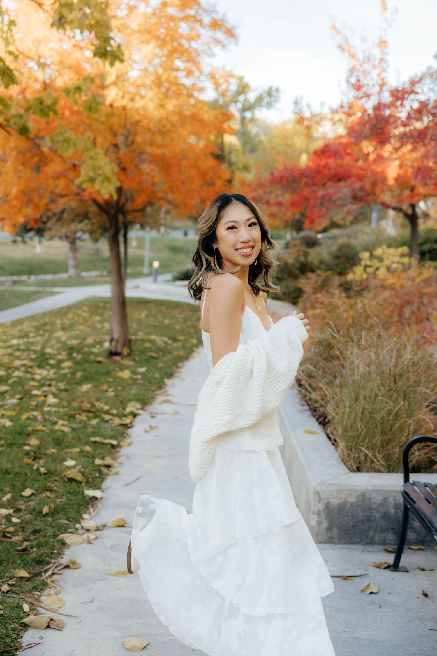 A white wedding dress twirls against a backdrop of vibrant autumn foliage and fallen leaves in a park setting.