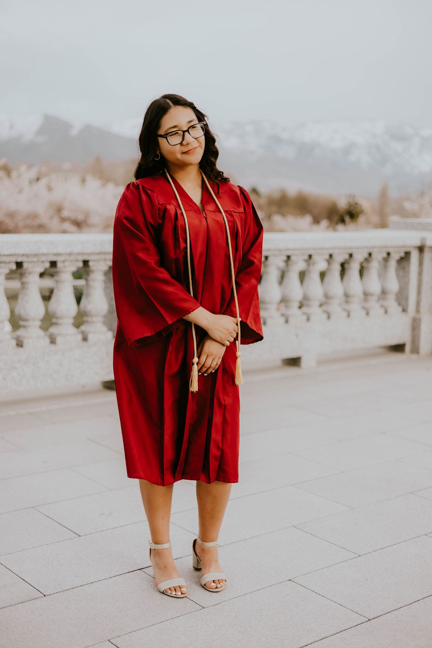 A graduate poses in a red graduation gown and silver heels on a stone terrace.