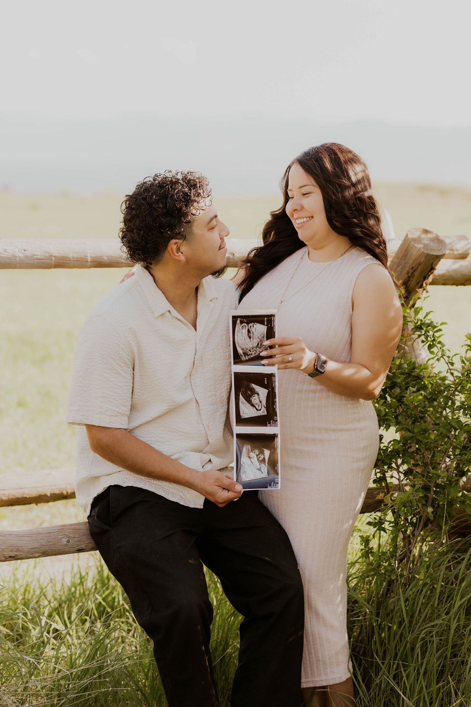 Couple sitting in grass field sharing ultrasound photos during golden hour photoshoot.