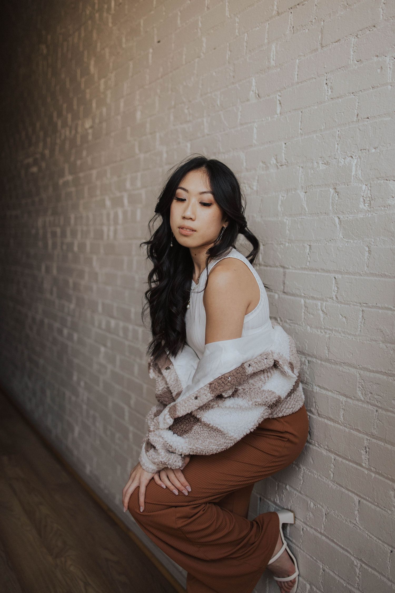 Moody portrait against white brick wall wearing cozy sweater and brown pants.