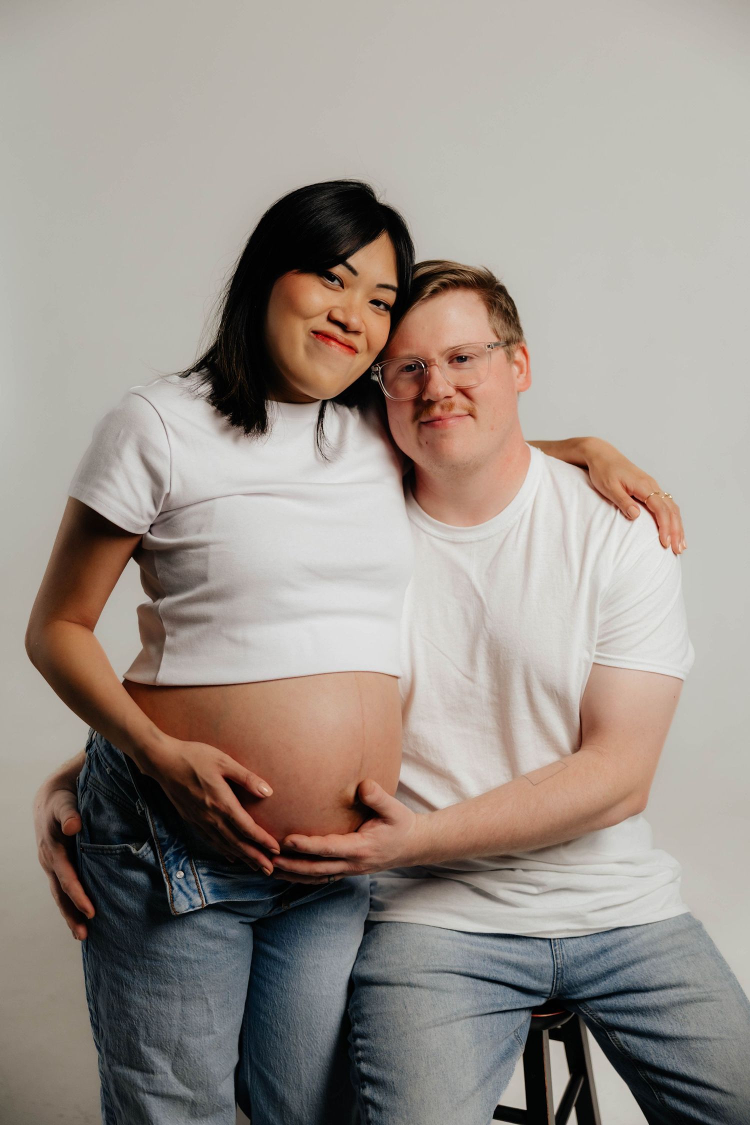 Intimate maternity photoshoot featuring couple in white shirts against neutral background.