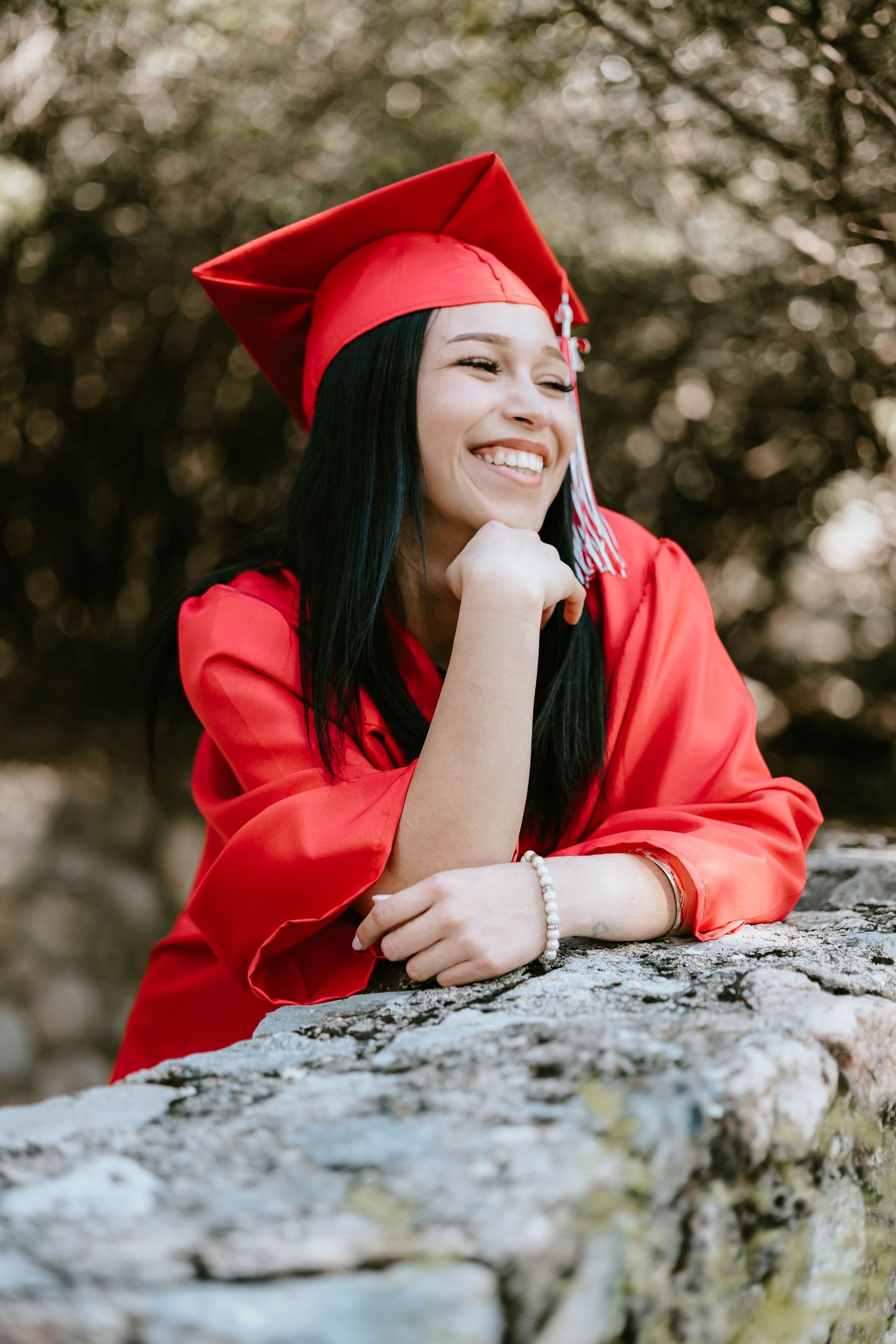 Graduate in red cap and gown poses playfully on a stone wall during outdoor photo session.
