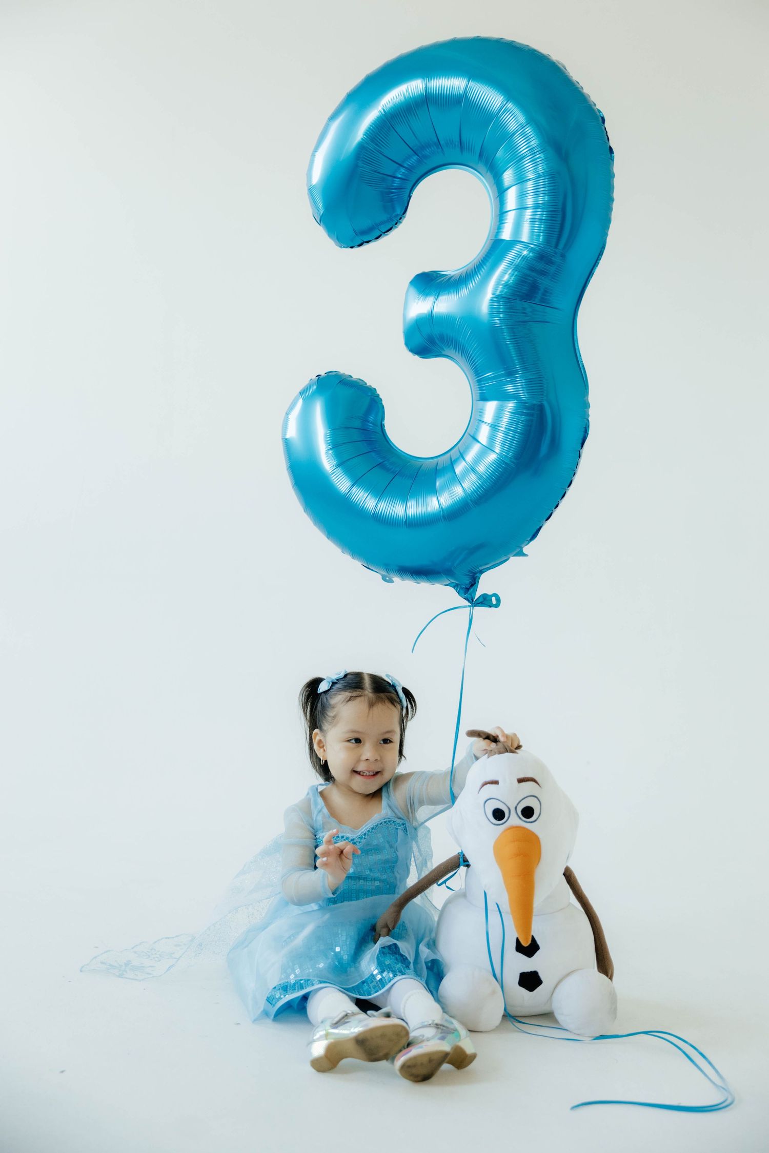Child in blue dress poses with Olaf plush toy and large blue number 3 balloon.