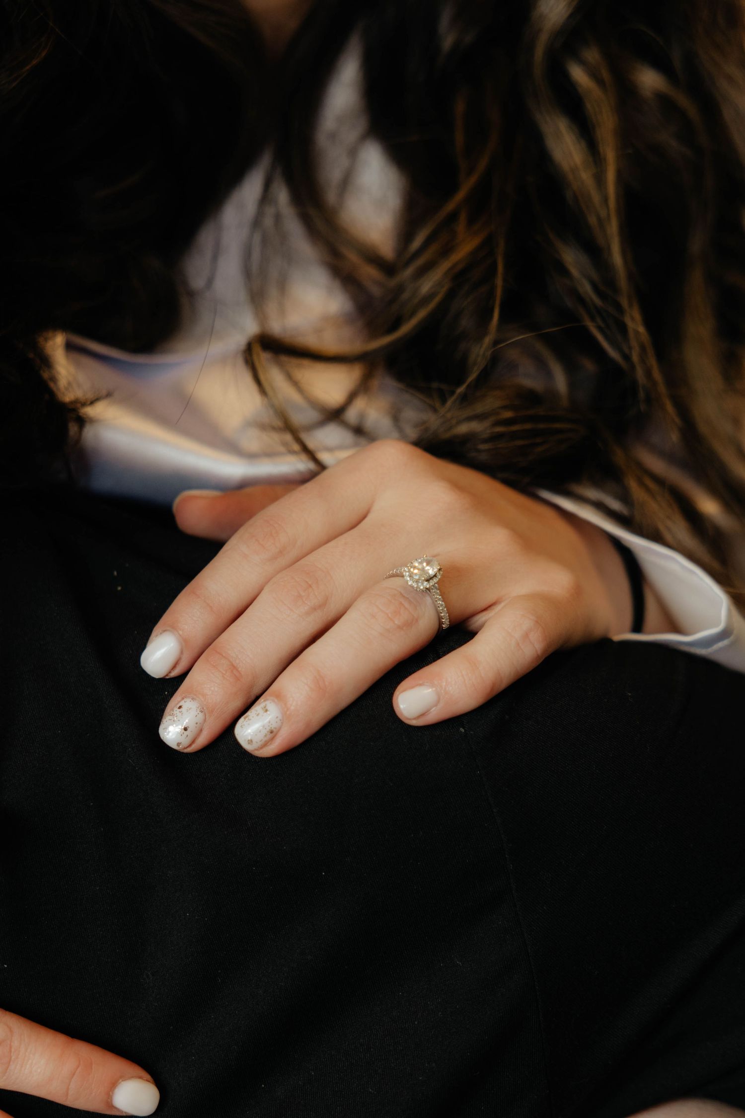 Close-up of hands showing off an engagement ring with white nail polish against a dark background.