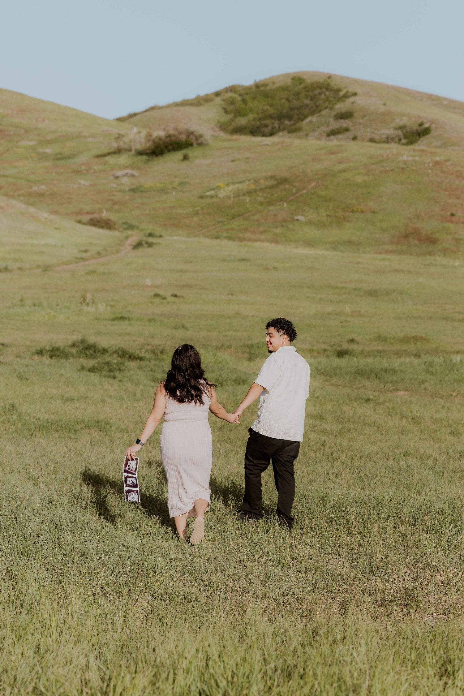Couple walking through grassy hillside holding ultrasound photos at sunset.