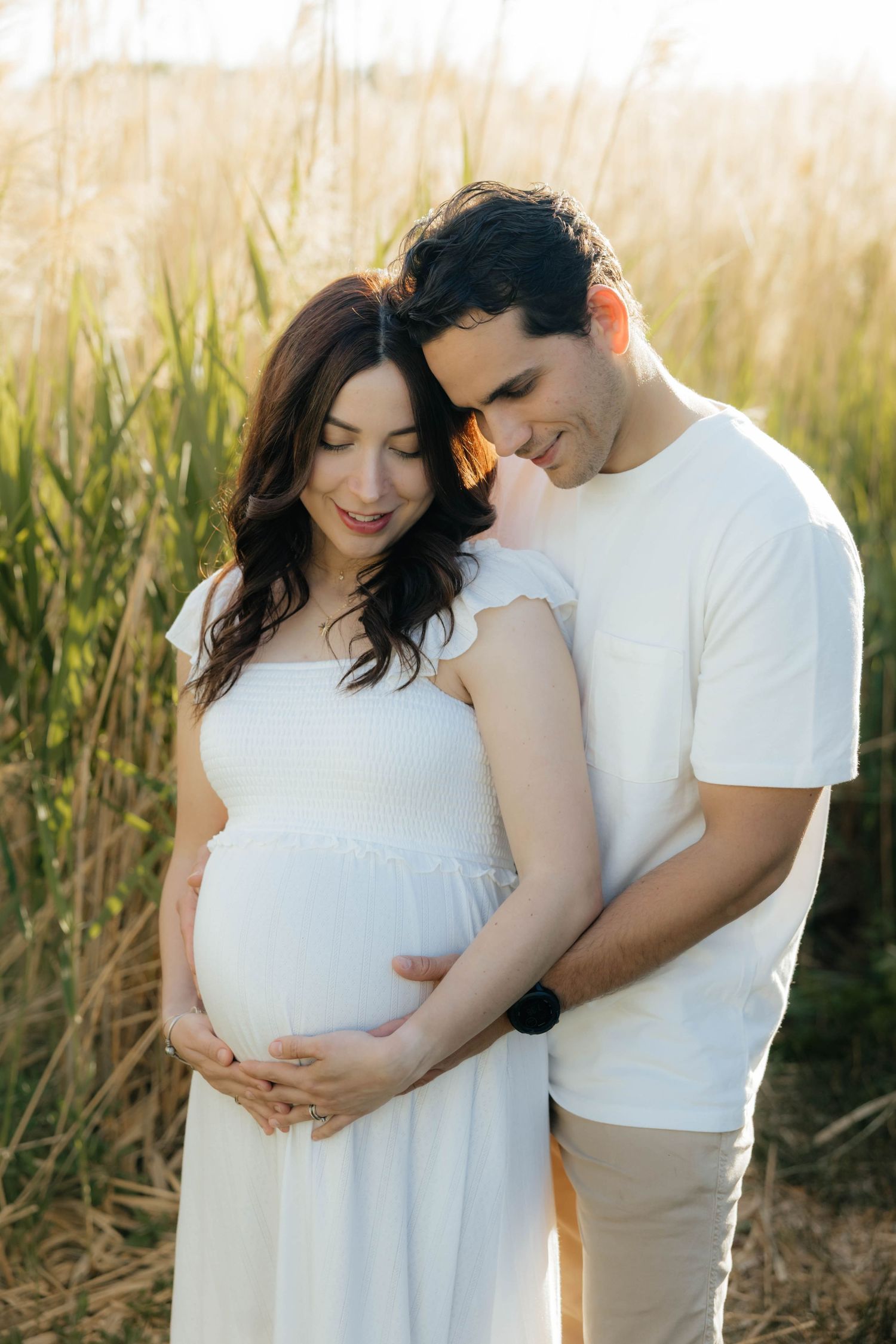 Intimate maternity photoshoot in a sunlit field with tall grass showing a couple in white clothing.
