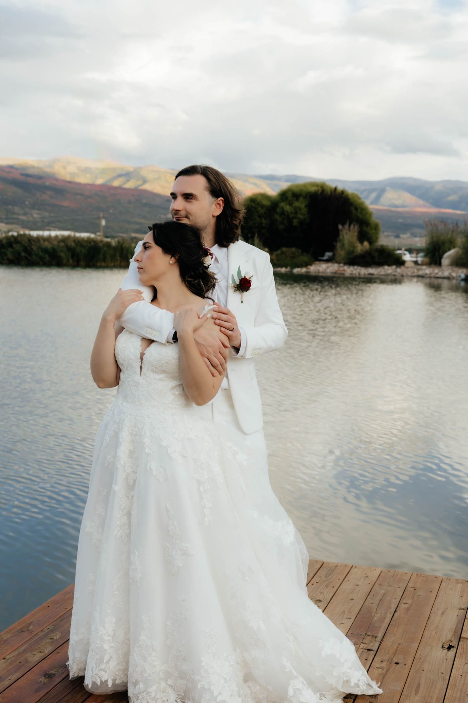 Couple in wedding attire shares intimate moment on dock by mountain lake.