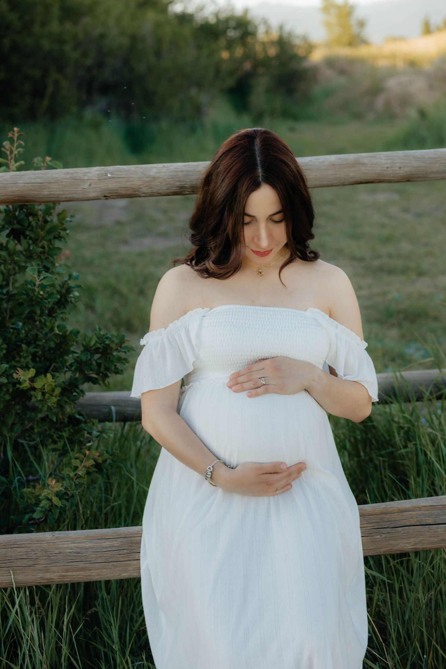 Outdoor maternity portraits featuring a woman in a flowing white off-shoulder dress by a wooden fence.