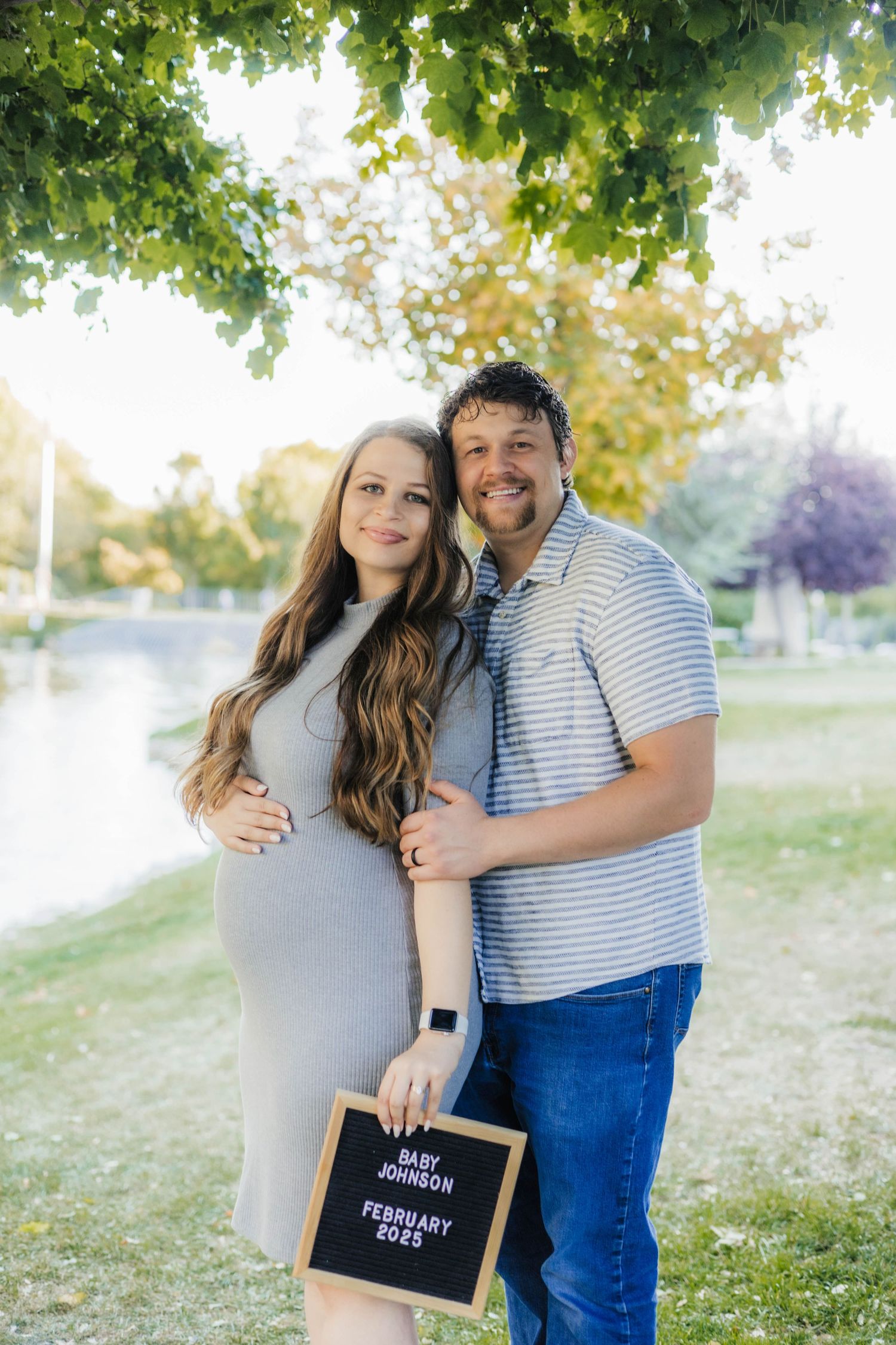 Couple poses outdoors holding a pregnancy announcement sign in a sunny park setting.