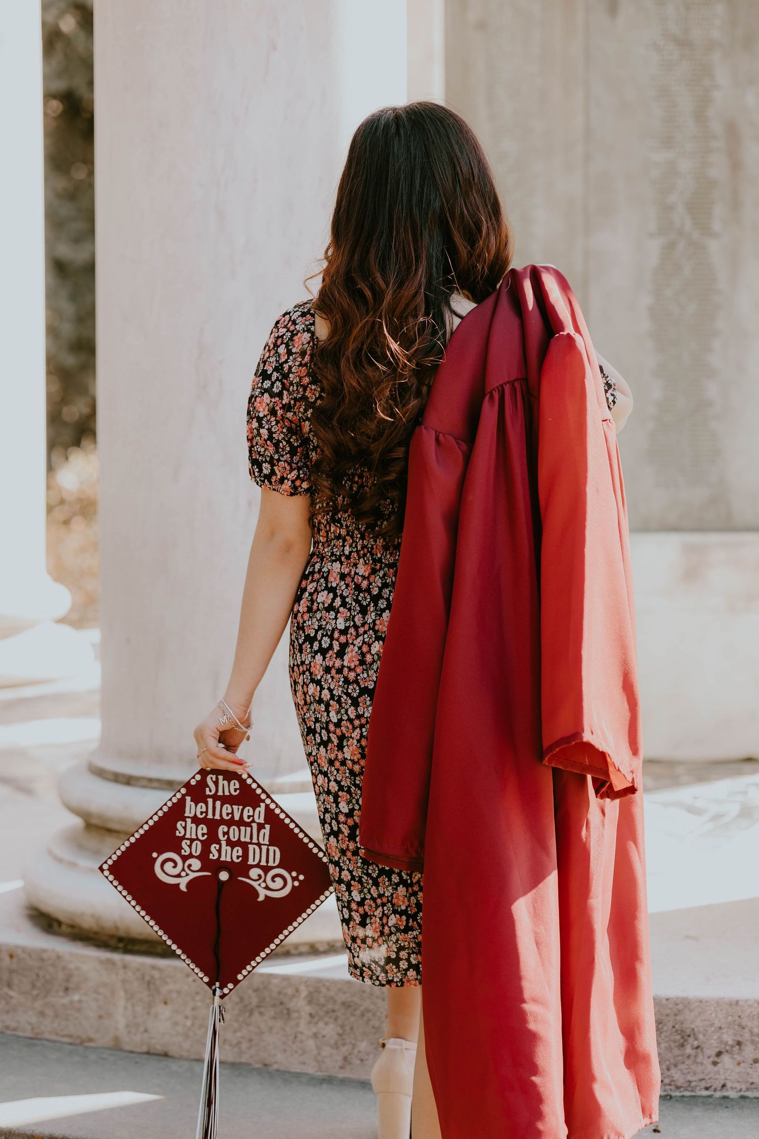 Person in floral dress holding red graduation cap outdoors.