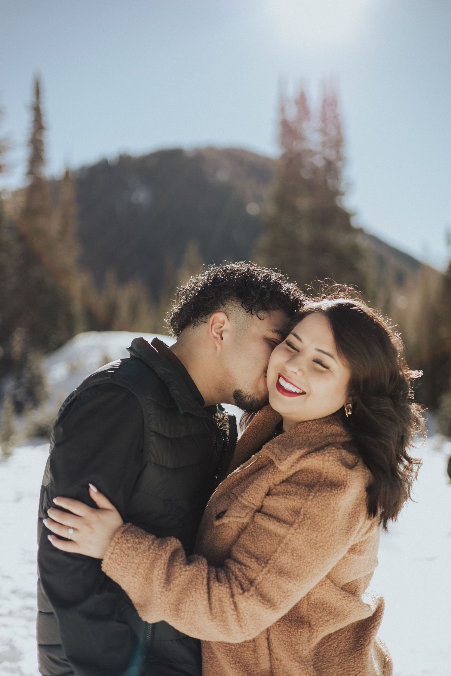 Couple sharing an intimate moment in winter with snow-capped mountains and evergreen trees in the background.