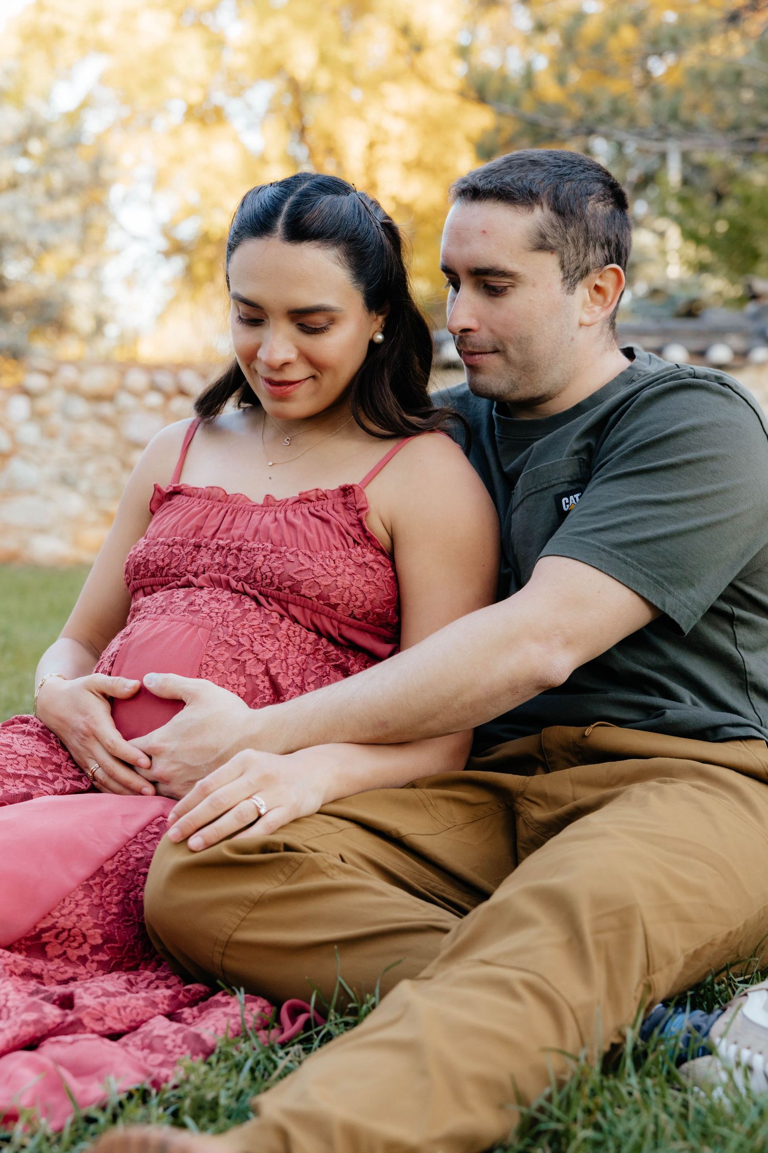 Couple lounging on grass during autumn maternity photoshoot wearing burgundy dress and casual attire.