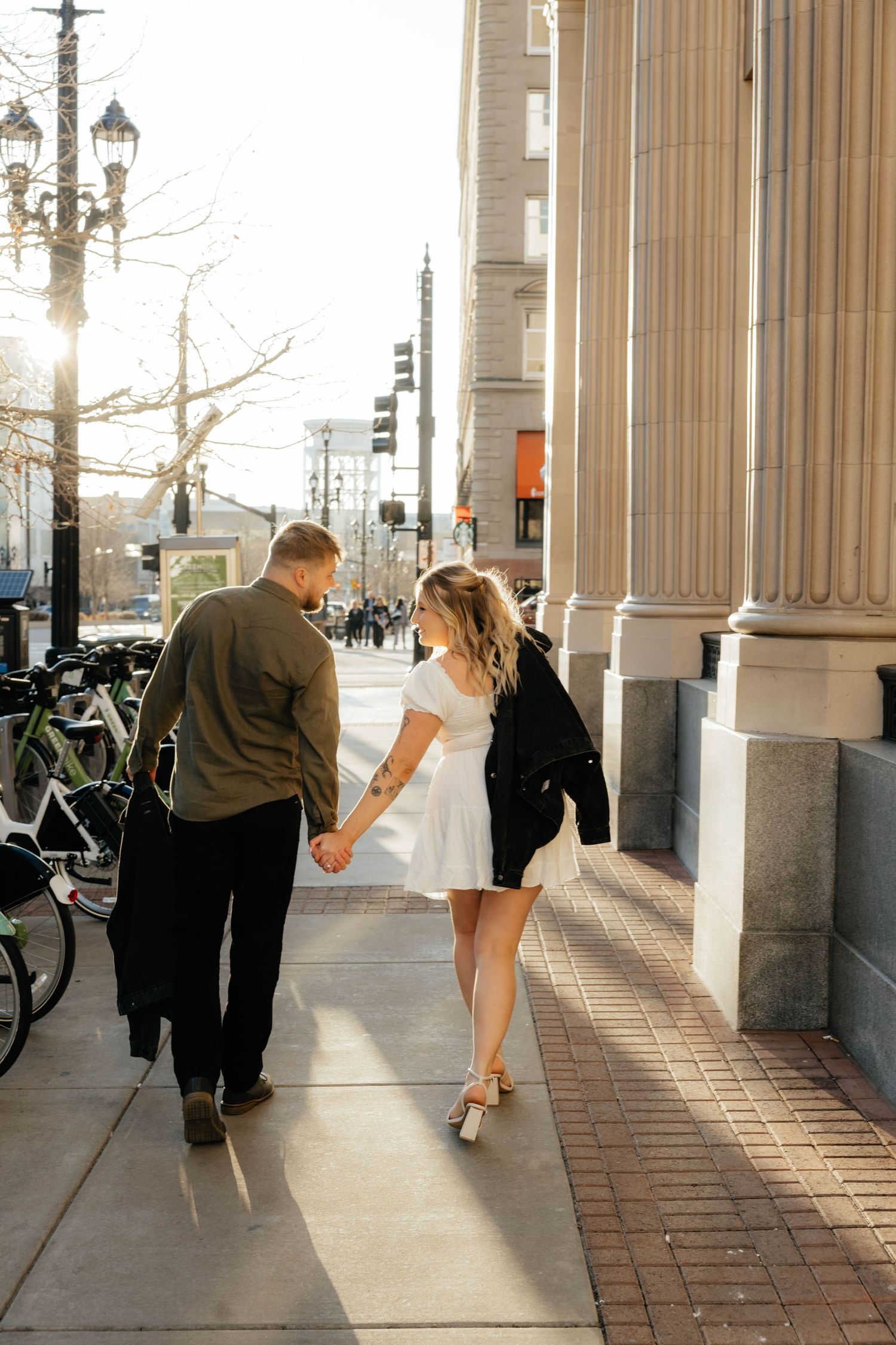 Playful couple dancing and spinning under classic stone columns on brick sidewalk.