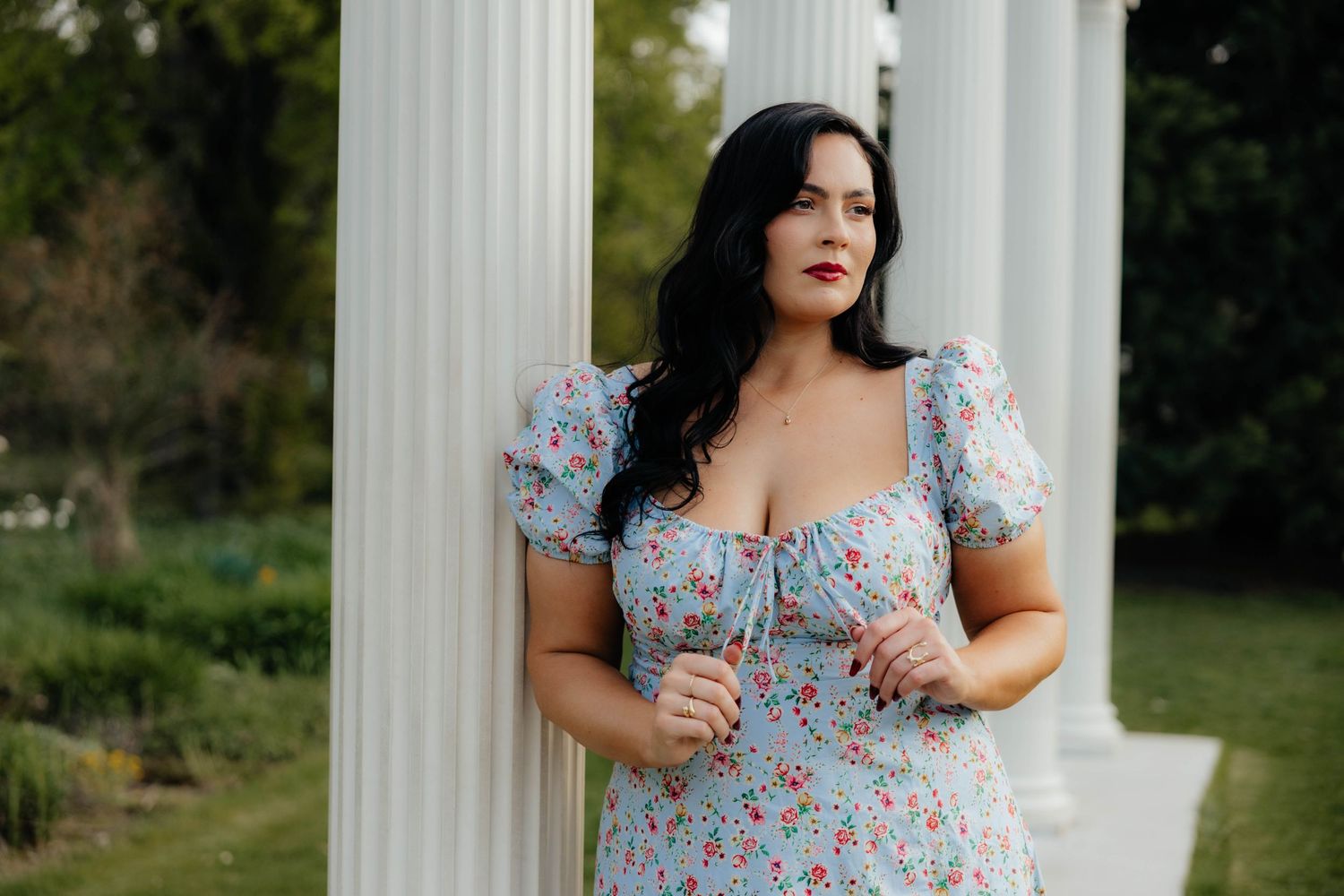 Person in floral blue dress poses against white classical columns outdoors.