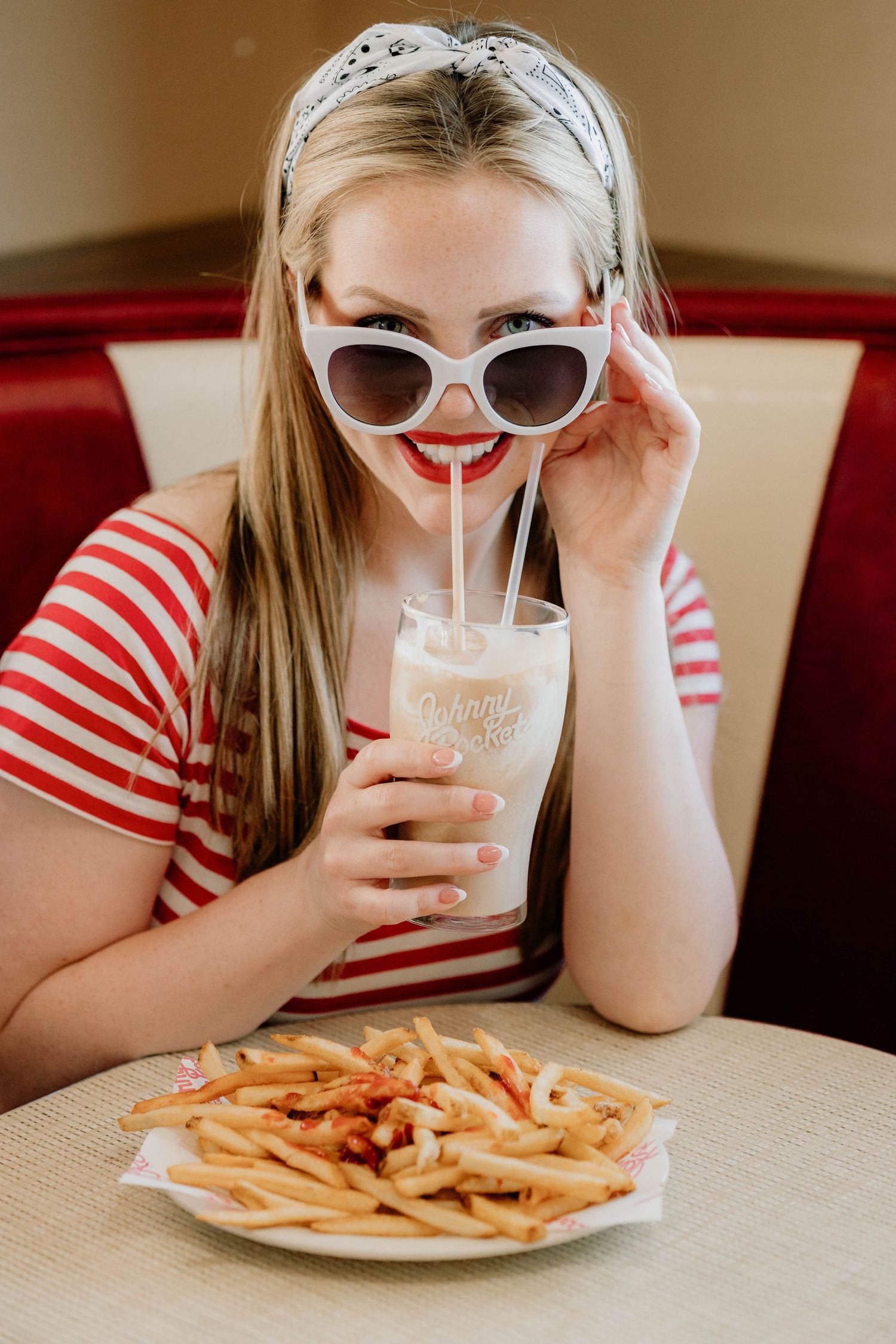 Person in striped shirt and sunglasses enjoying a milkshake and french fries at a diner table.