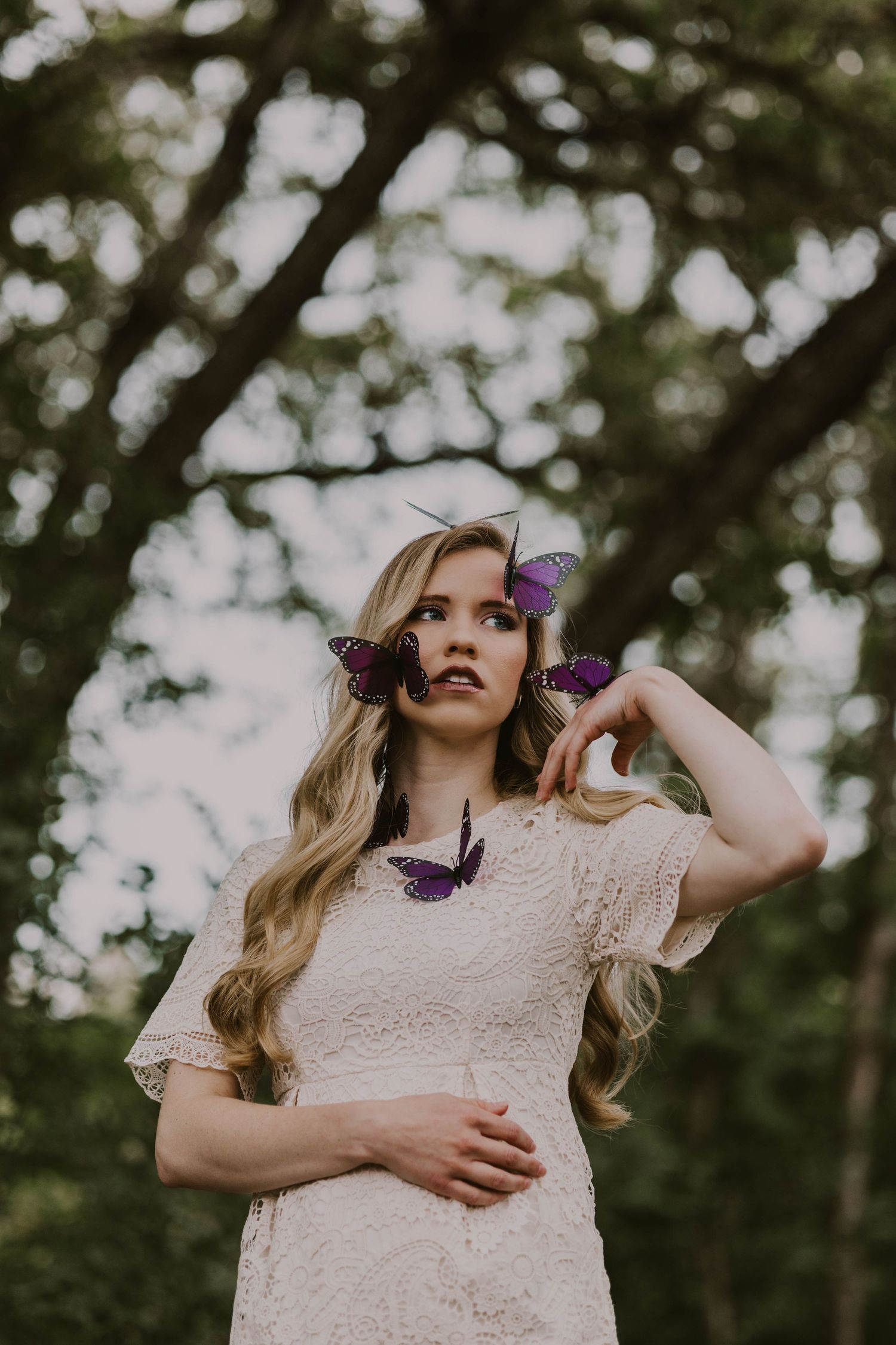Whimsical outdoor portrait with purple butterflies and lace dress against blurred trees.