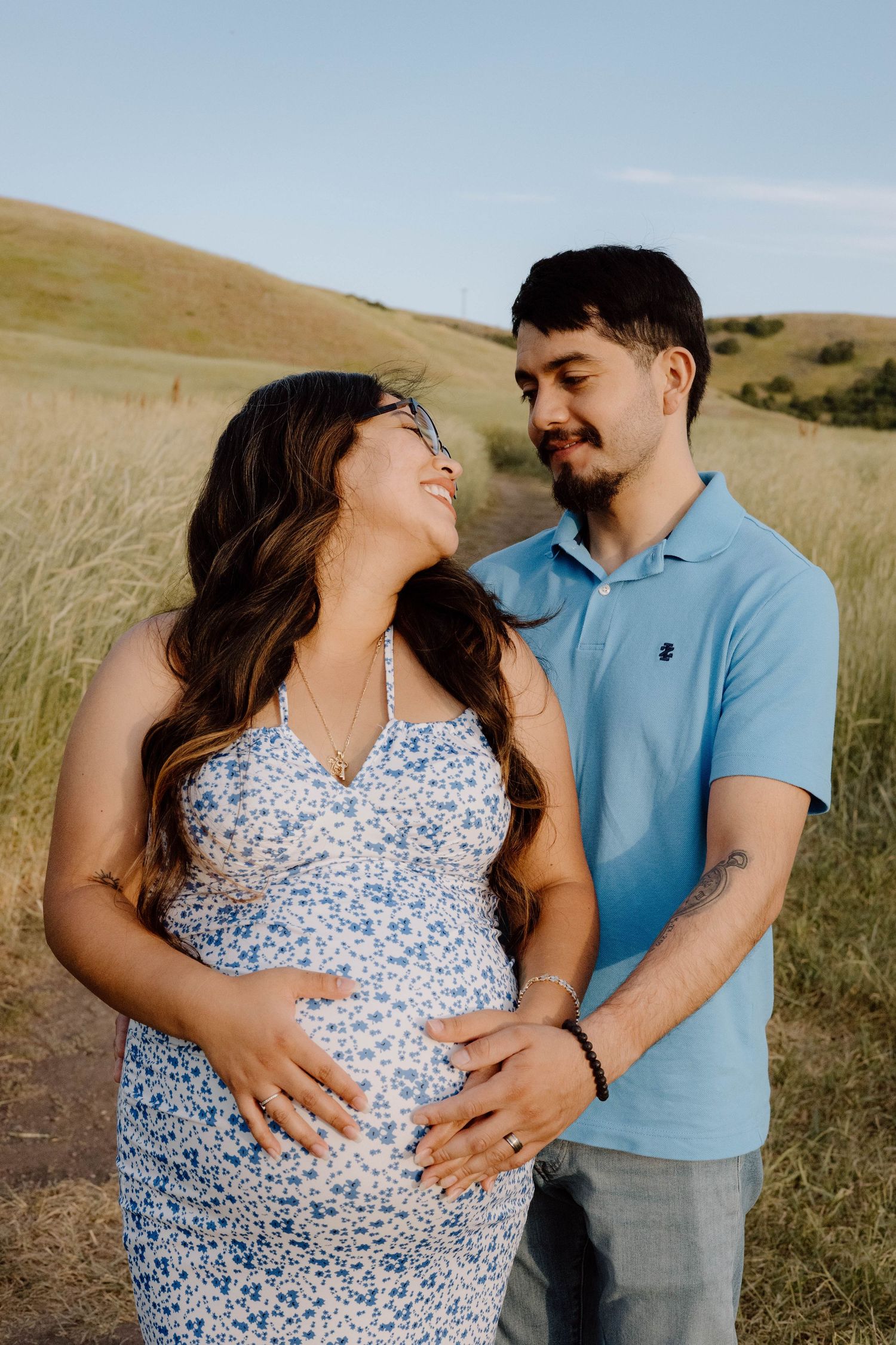 A couple poses together in a grassy field during a maternity photoshoot at golden hour.