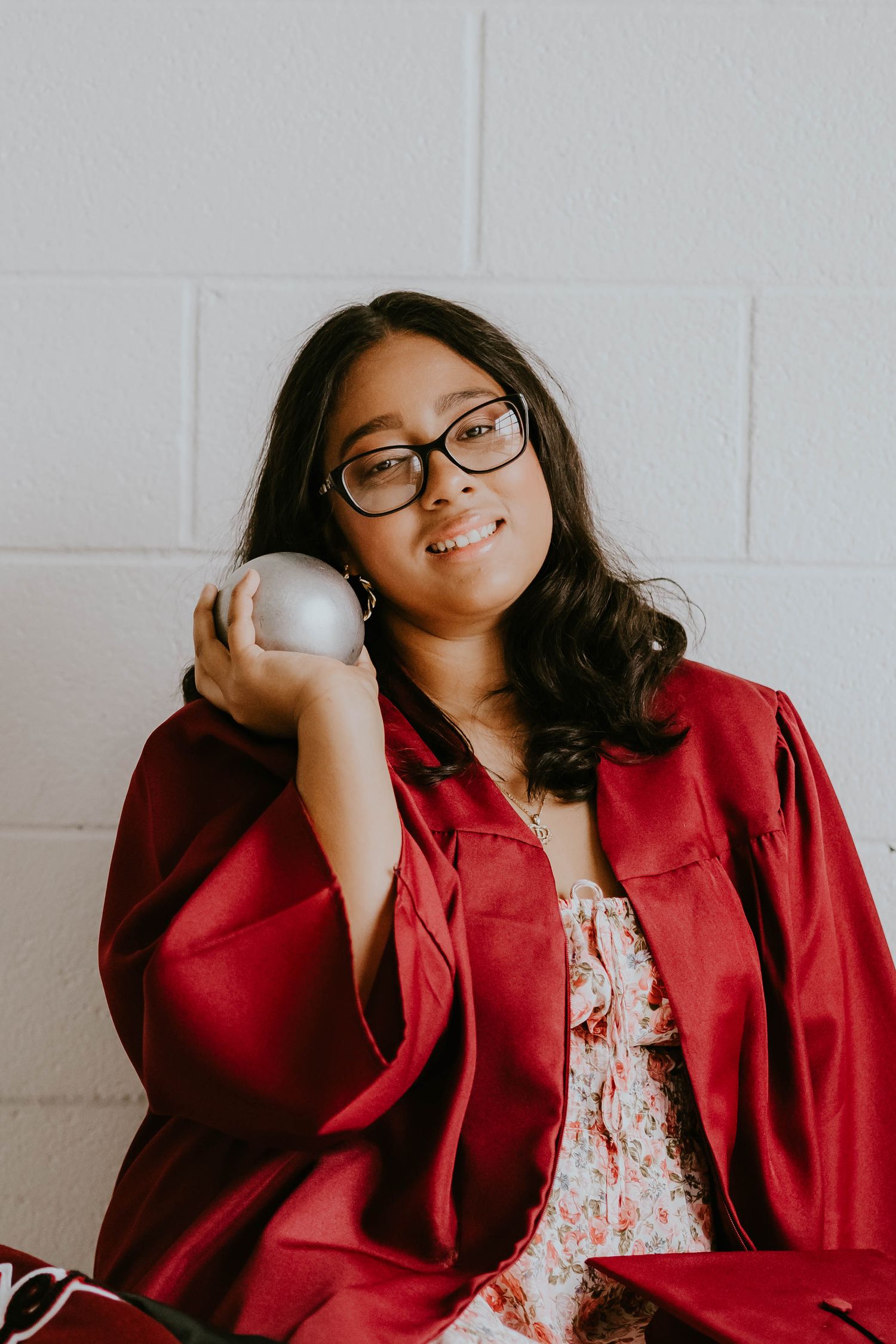 Graduate in red gown poses against white brick wall during indoor photo session.