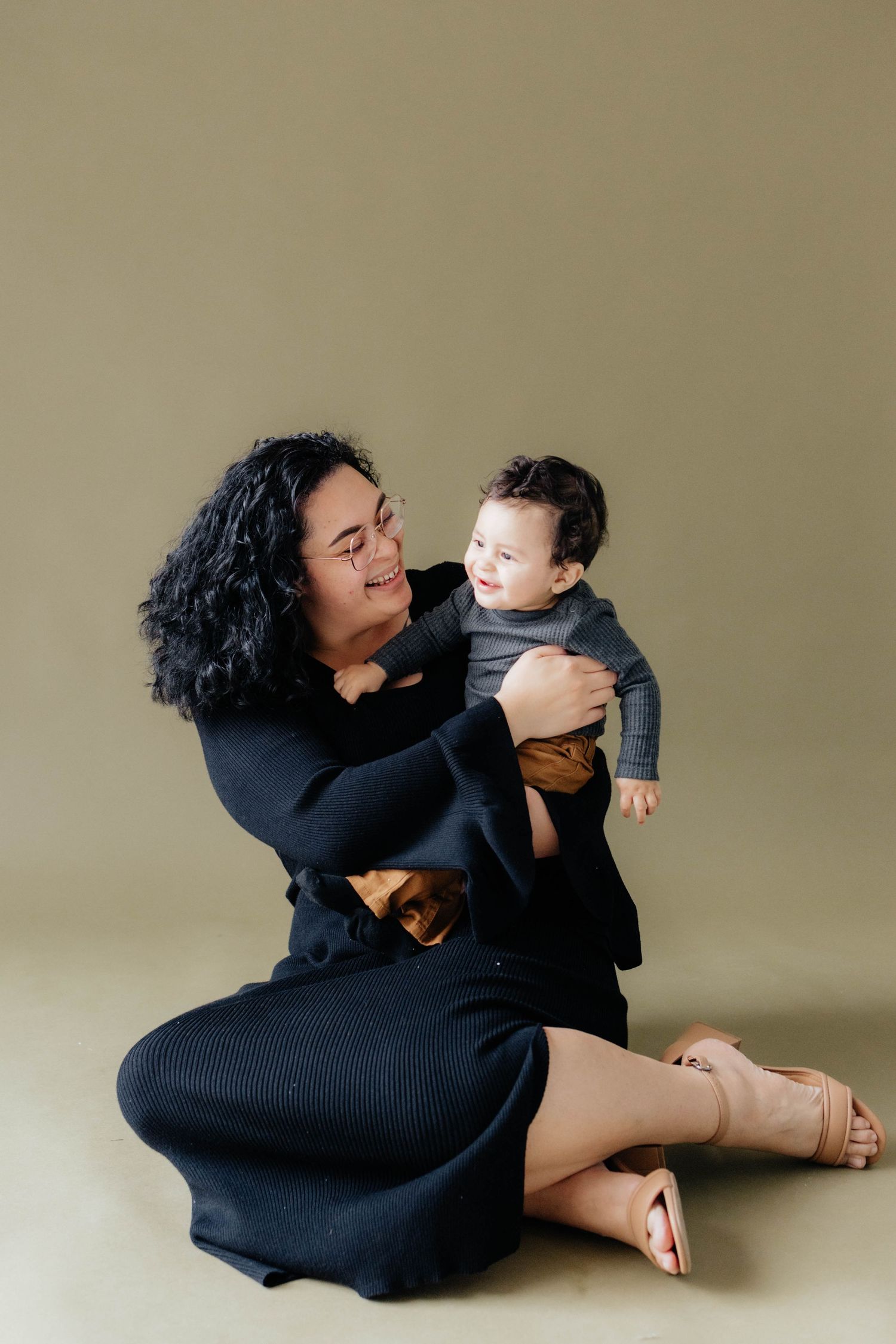 Parent in black dress sitting on floor lovingly holding and looking at baby in grey outfit.