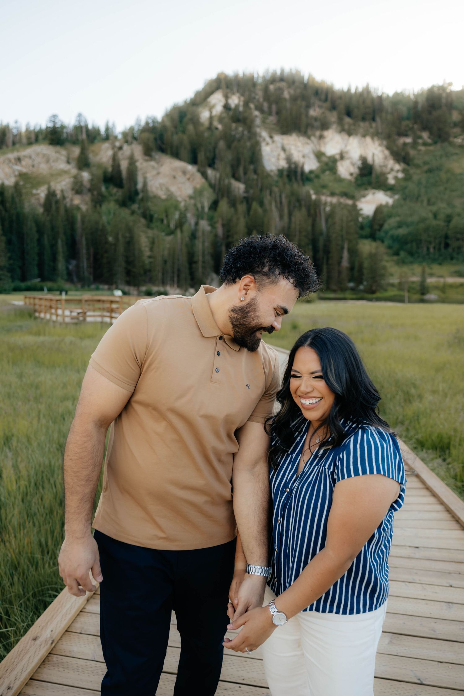 Couple sharing intimate laughs on wooden boardwalk with mountain landscape backdrop.