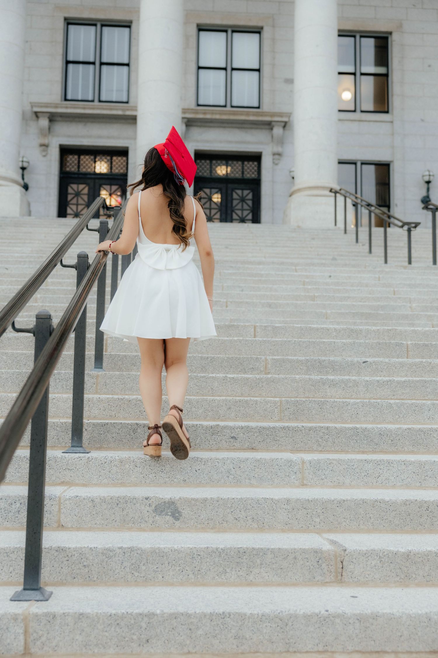 Graduate in white dress and red cap ascends stone steps of academic building.