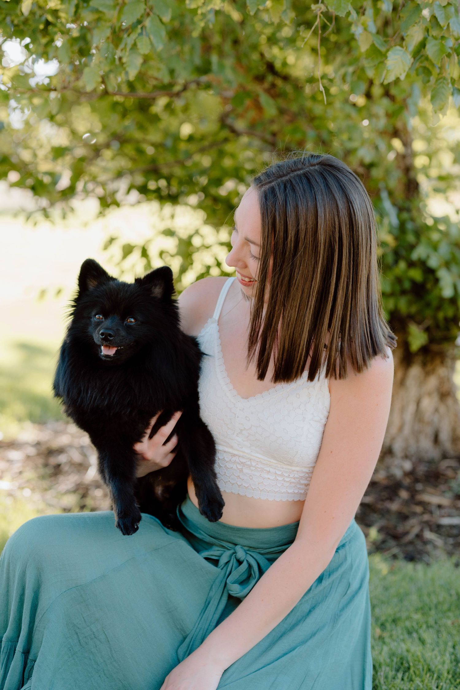 A candid moment showing a person in teal skirt holding a fluffy black Pomeranian dog outdoors.