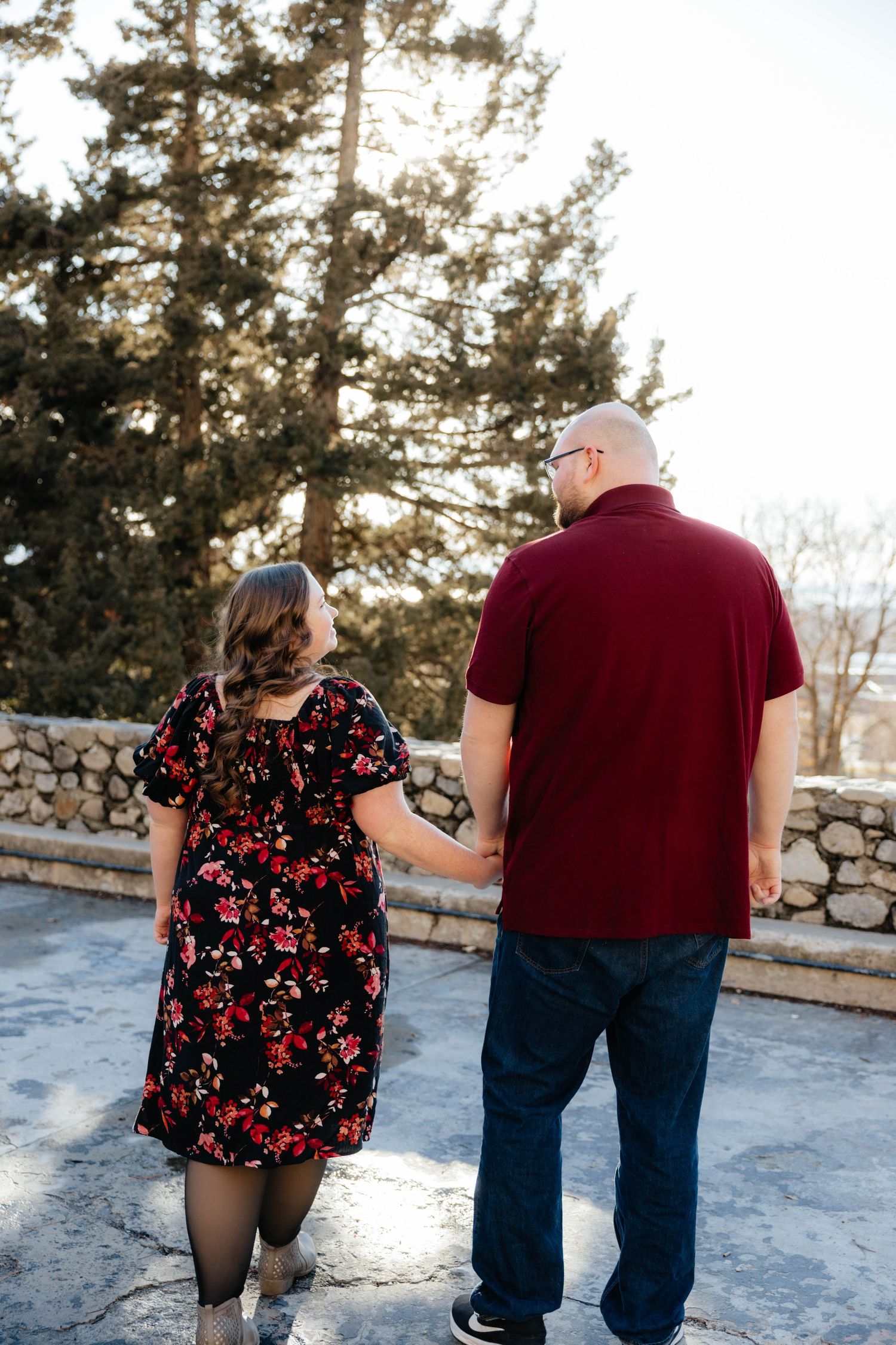 Playful couple walking in the snow with stone wall and pine trees in background.
