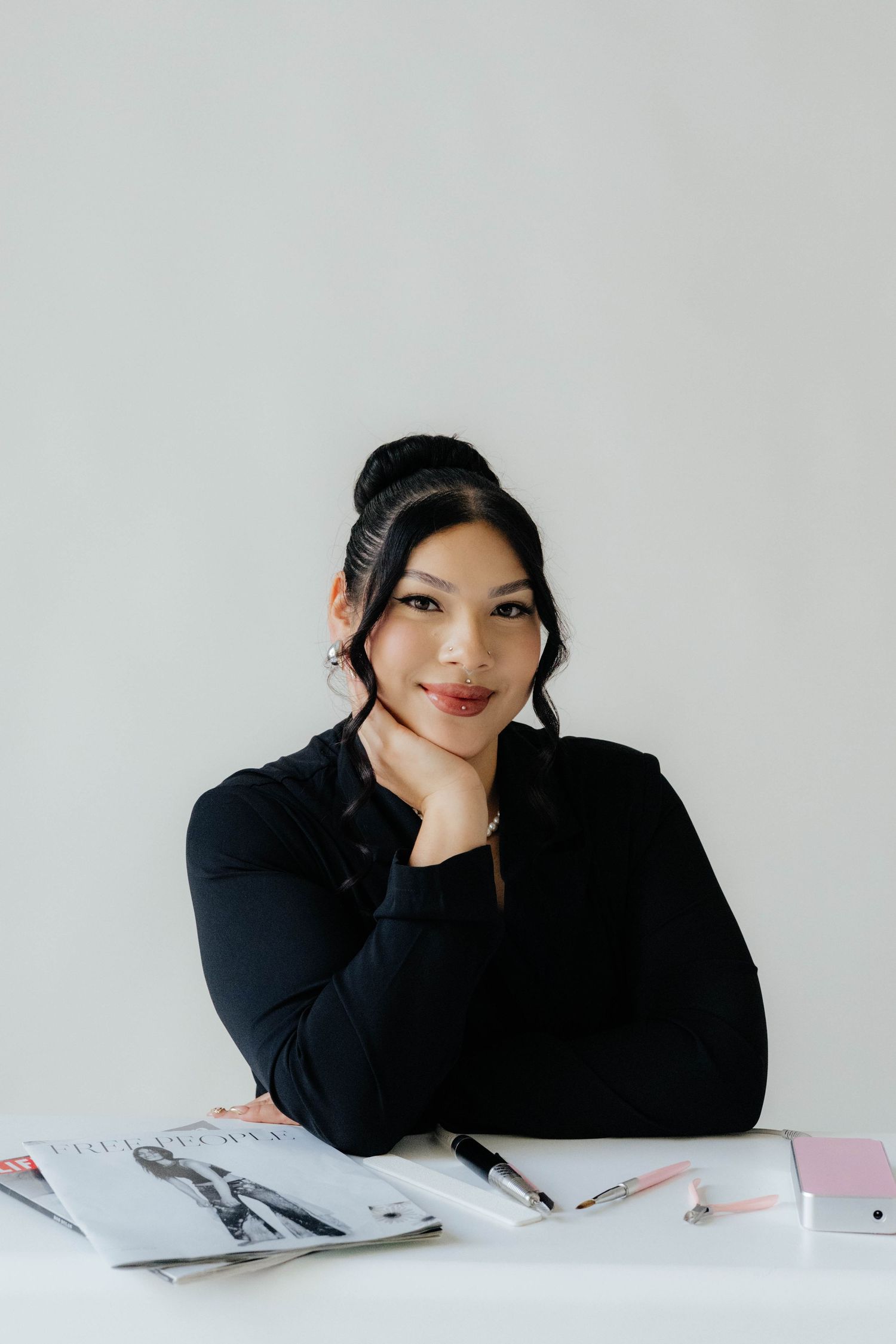 Professional in black outfit sits at desk with magazine and writing implements.